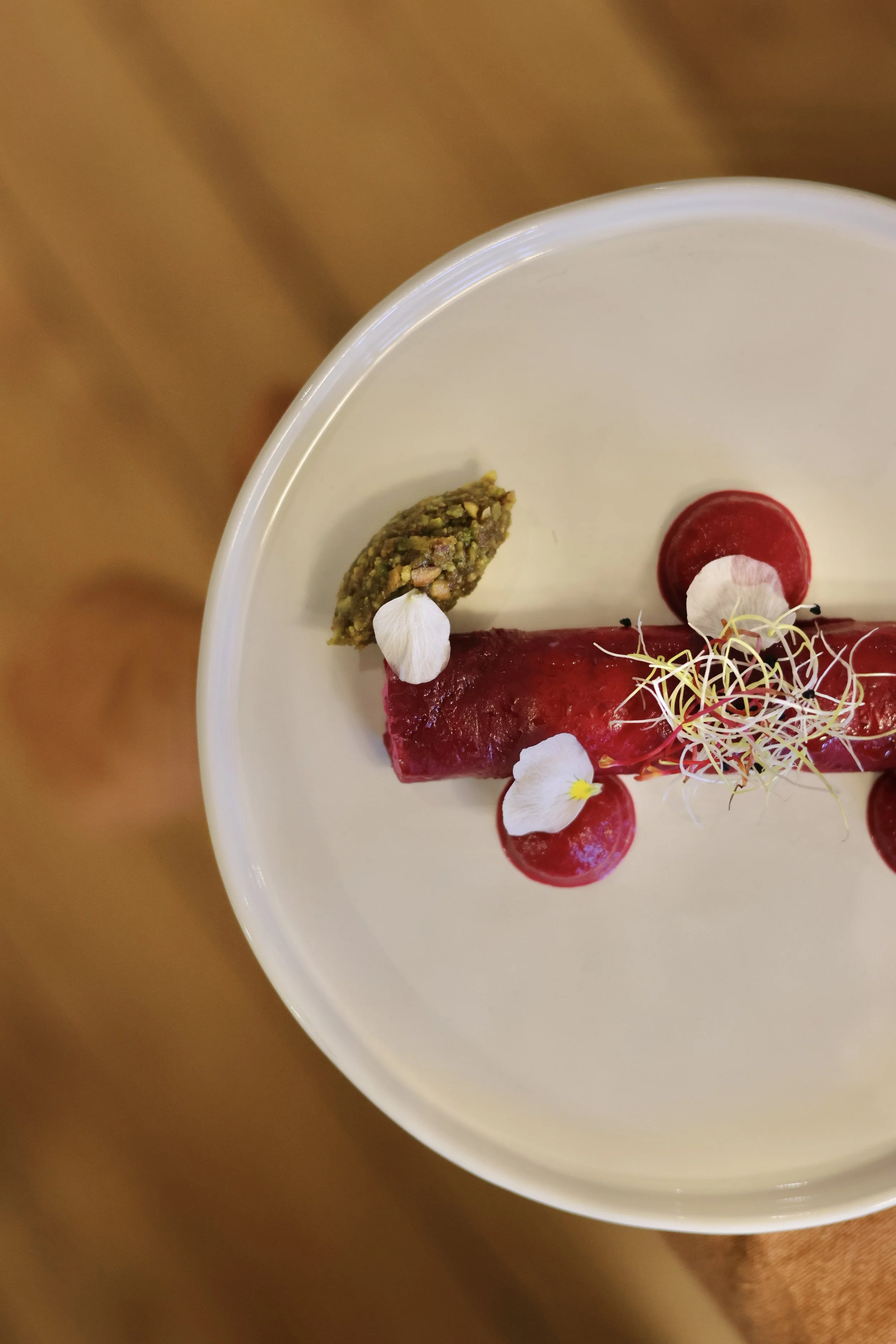 Un plat de dessert avec une fine couche de gelée rouge, une petite brochette de pistaches hachées et une garniture de fleurs comestibles et de jeunes pousses.