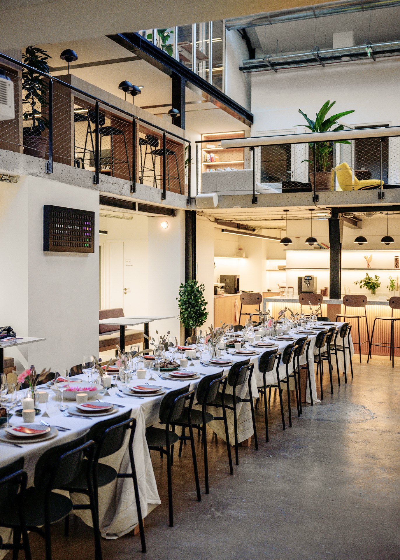Une salle de réception élégante avec une grande table dressée pour un repas, décorée de vaisselle, de verres et de bouquets de fleurs. La salle a un design industriel avec un mélange de mobilier moderne et quelques plantes vertes. Un espace bar ou ki