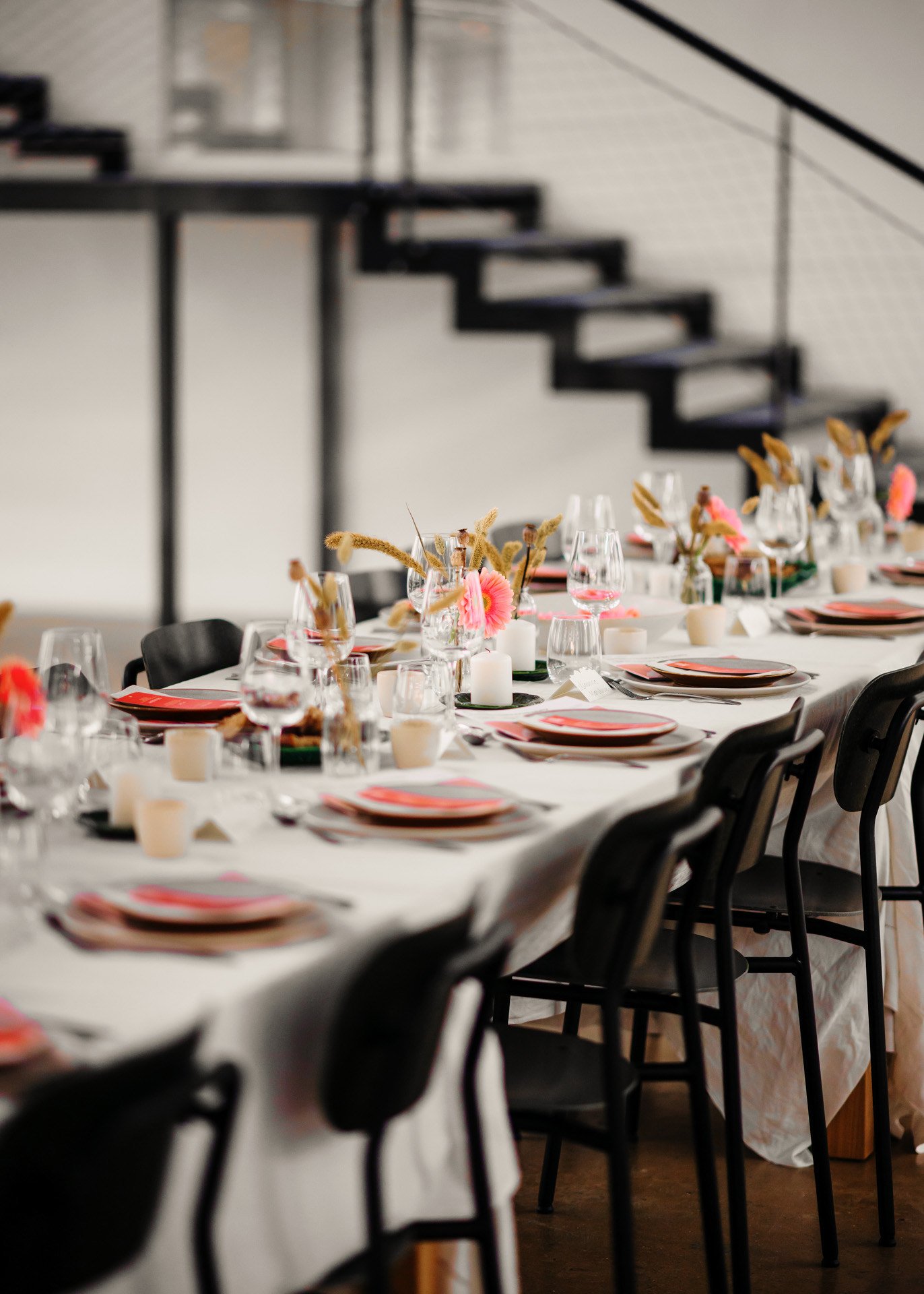 une longue table dressée pour un repas, avec des assiettes, des verres, des couverts, des bougies et des arrangements floraux, dans un espace intérieur moderne.