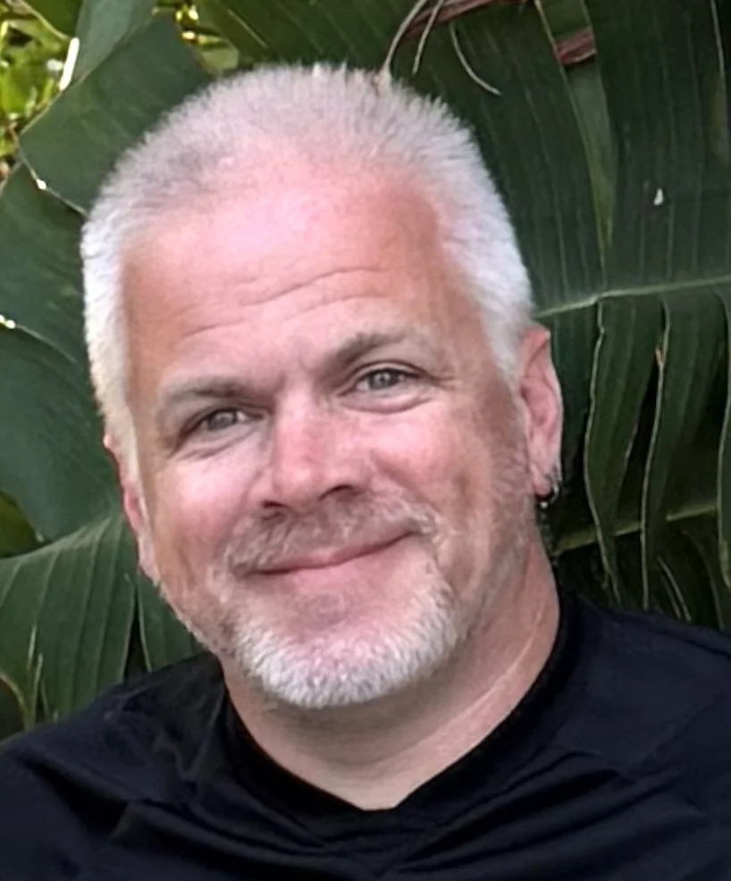 Close-up of a smiling man with short white hair, a beard, and an earring, standing outdoors in front of green tropical plants.