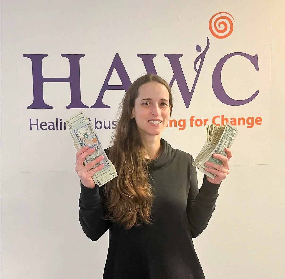 A woman with long brown hair holding up cash in both hands, standing in front of a sign 'HAWC Healing Abuse Working for Change'