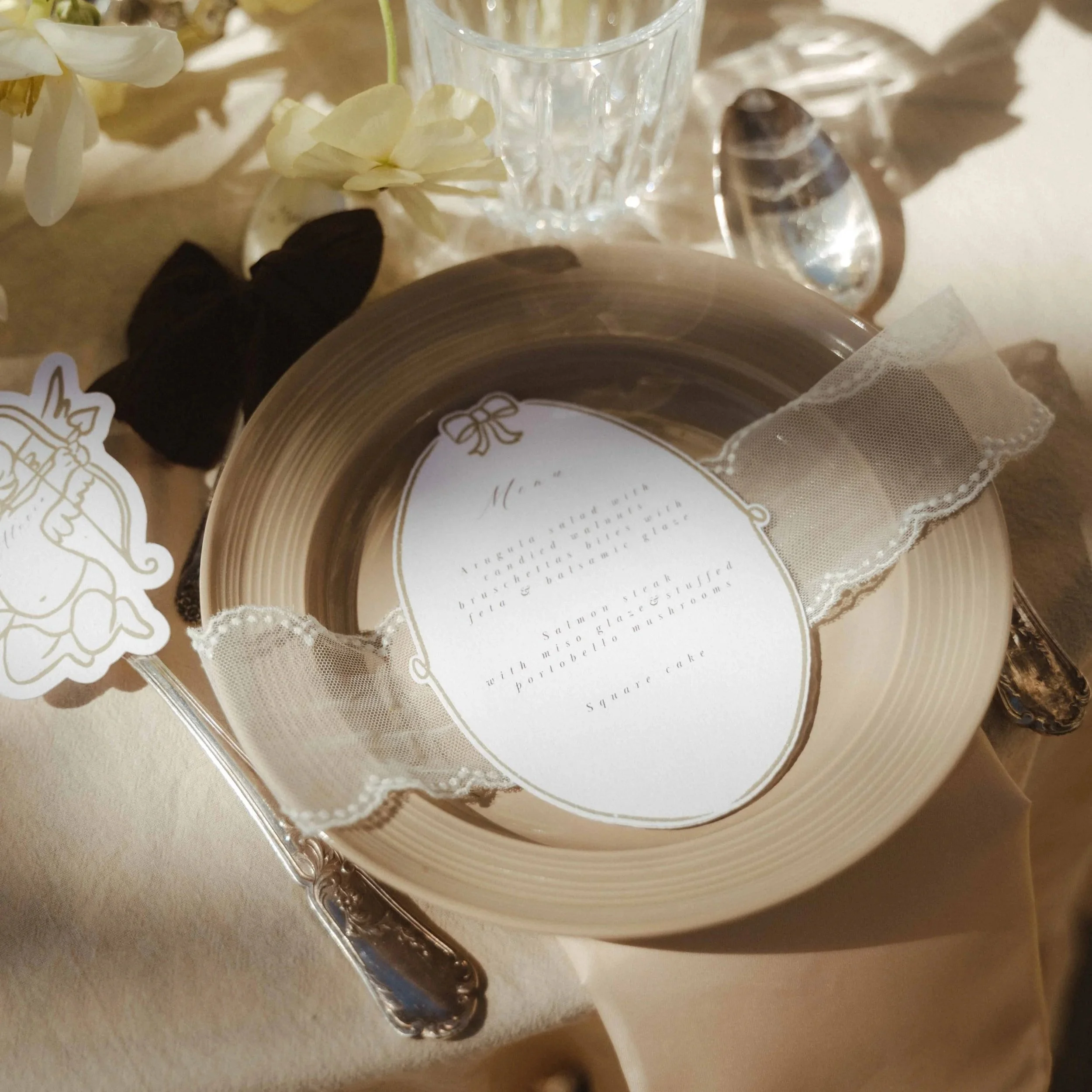 Décor de fête comprenant un verre en cristal, des fleurs blanches, des rubans, une étiquette de gâteau et des éléments décoratifs en dentelle, posés sur une surface beige.