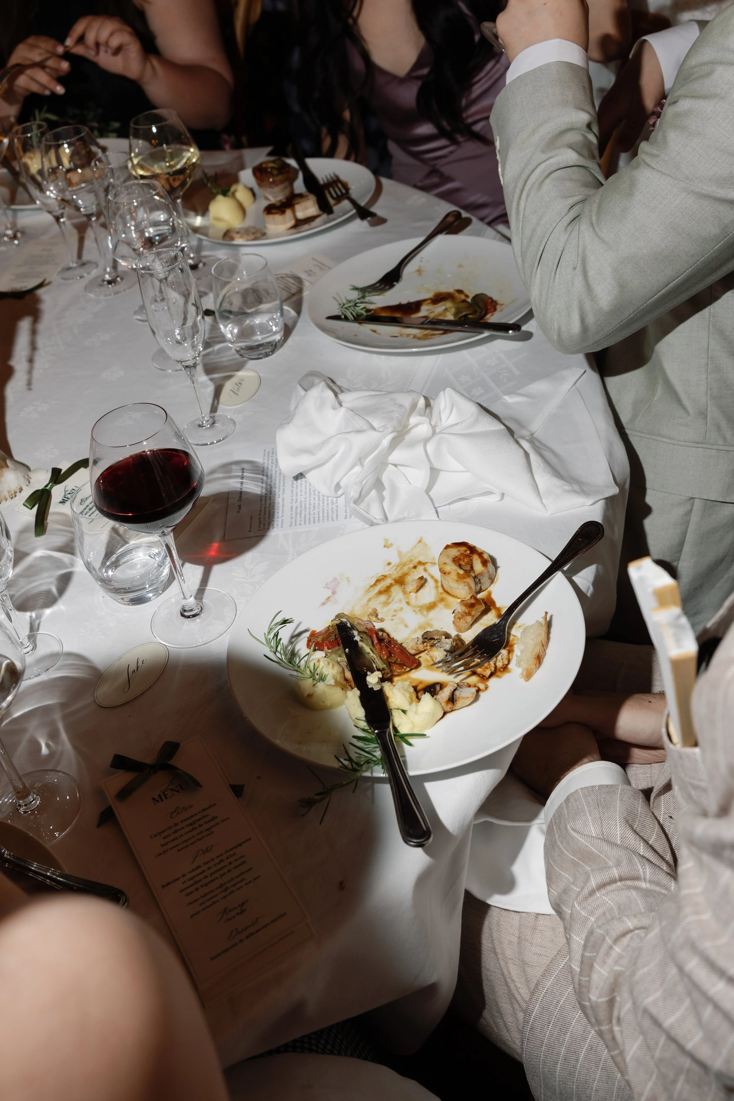 Table de dîner avec des assiettes vides et des verres de vin rouge, de l'eau et du champagne, des fourchettes, un menu et des restes de nourriture, entourée de personnes en costume, dans un cadre de repas formel.