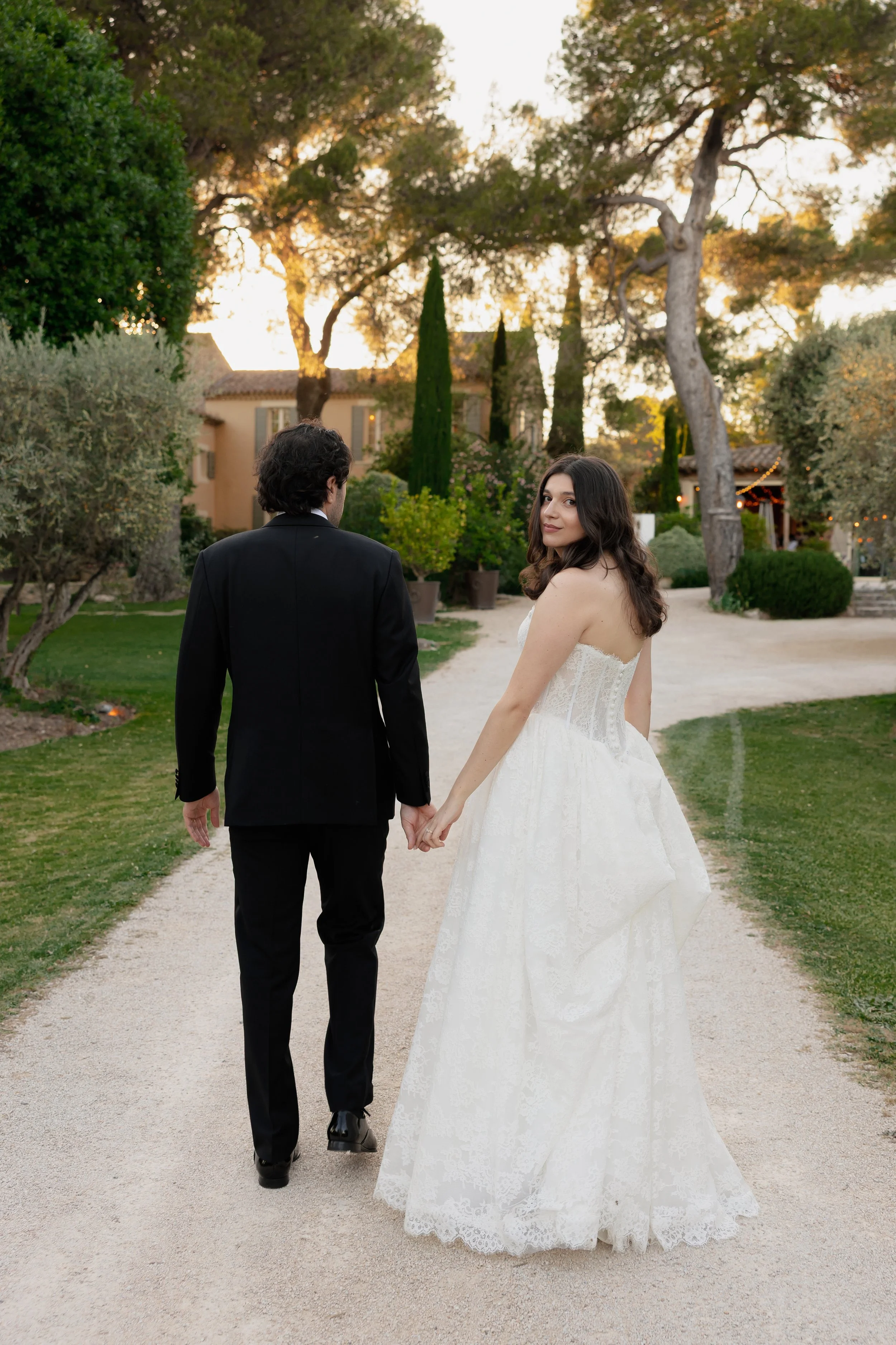 Un couple en mariage, la femme en robe blanche de mariage, l'homme en costume noir, marche dans un jardin, le coucher du soleil en arrière-plan.