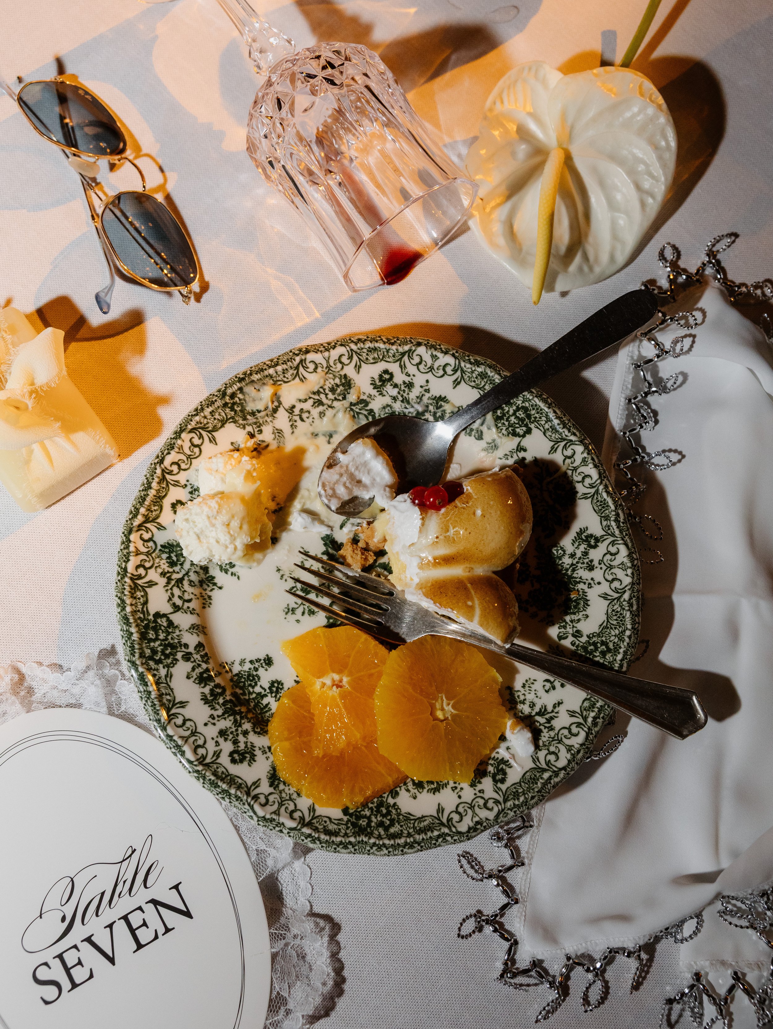 Un plateau avec des tranches d'orange, un gâteau à la crème et une tarte aux pommes, avec un verre à dessert à moitié rempli de sirop ou de vin rouge, une paire de lunettes de soleil et des bijoux argentés, le tout sur une nappe blanche avec une décoration en dentelle.