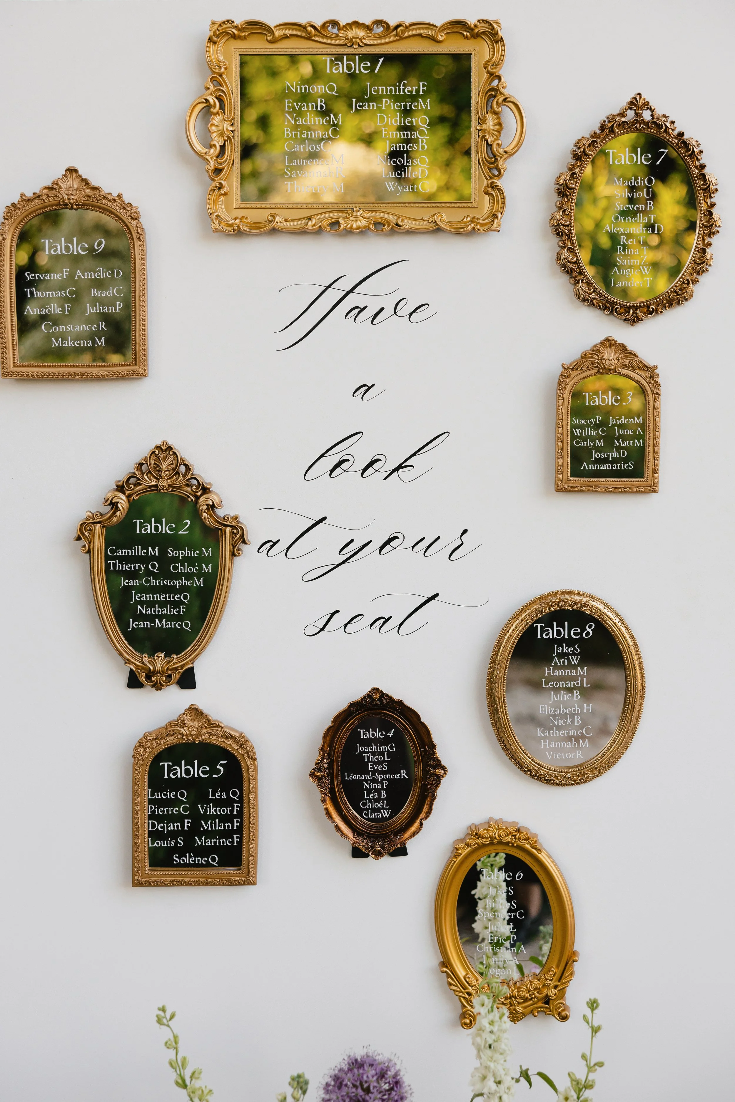 Tableau de mariage avec noms des invités sur des cadres dorés suspendus sur un mur blanc, décor floral en dessous.