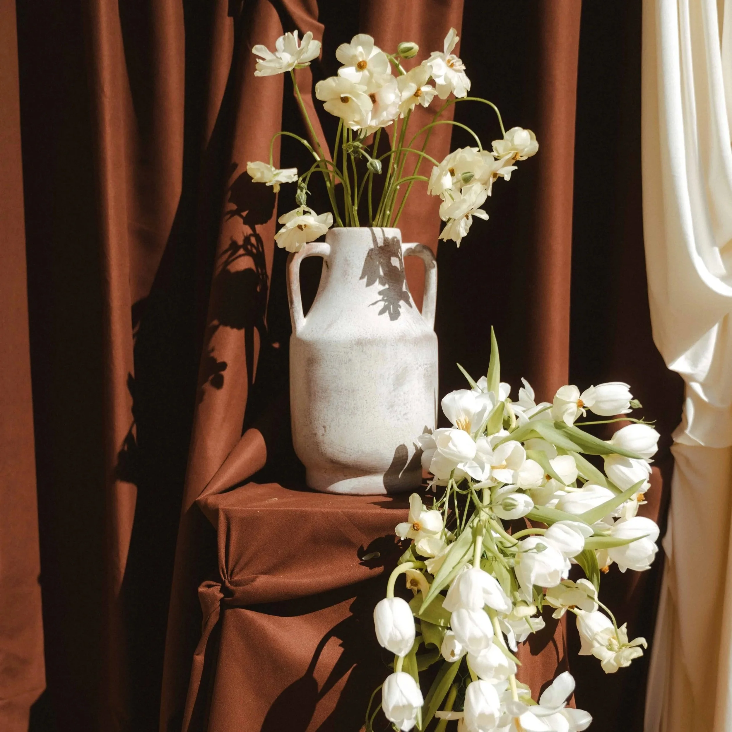 Un vase en céramique blanc contenant un bouquet de fleurs blanches, placé devant un rideau marron foncé et une autre de couleur crème.