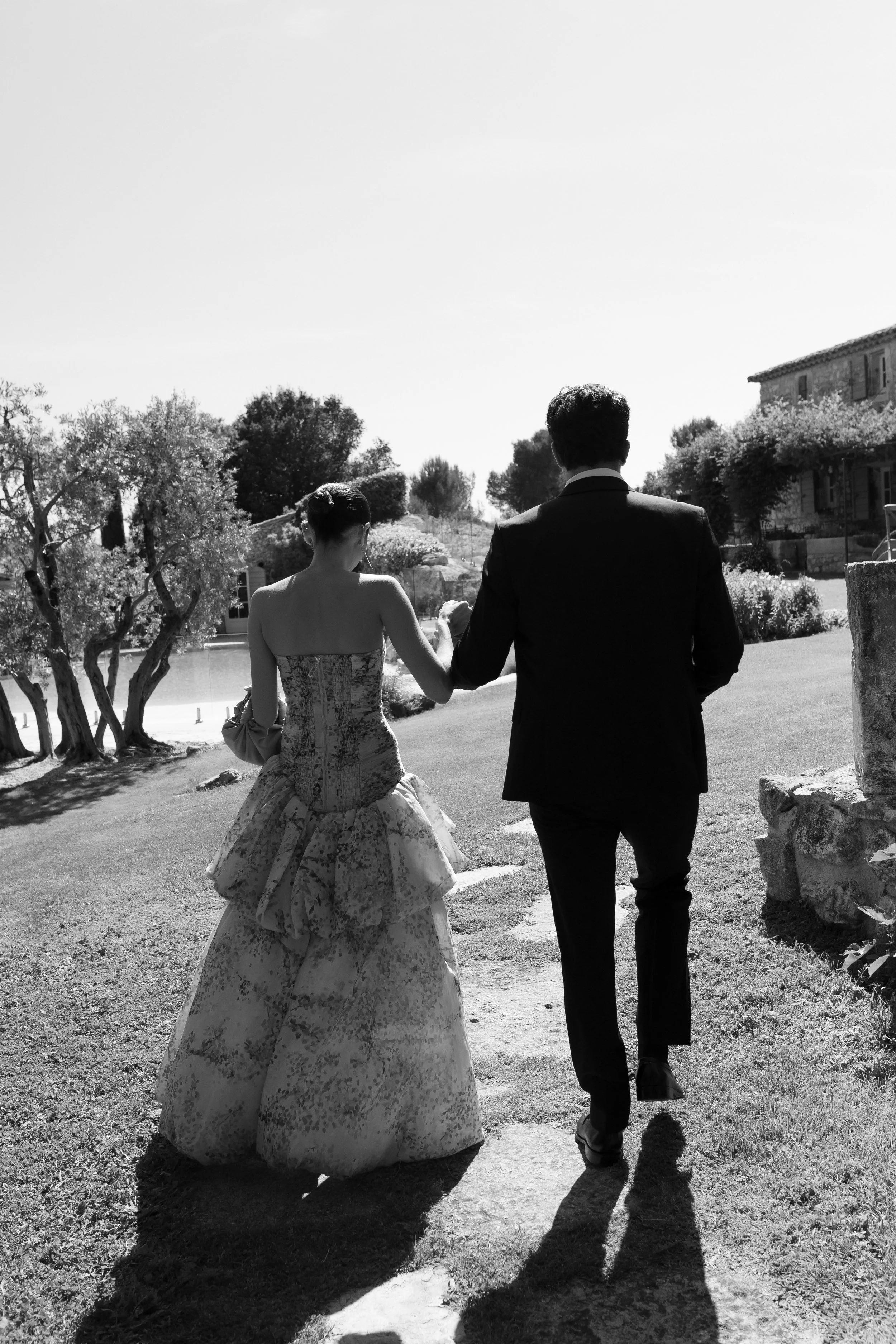 Photo en noir et blanc de deux personnes, un homme en costume et une femme en robe longue, marchant dans un jardin ensoleillé avec des arbres et une maison en arrière-plan.