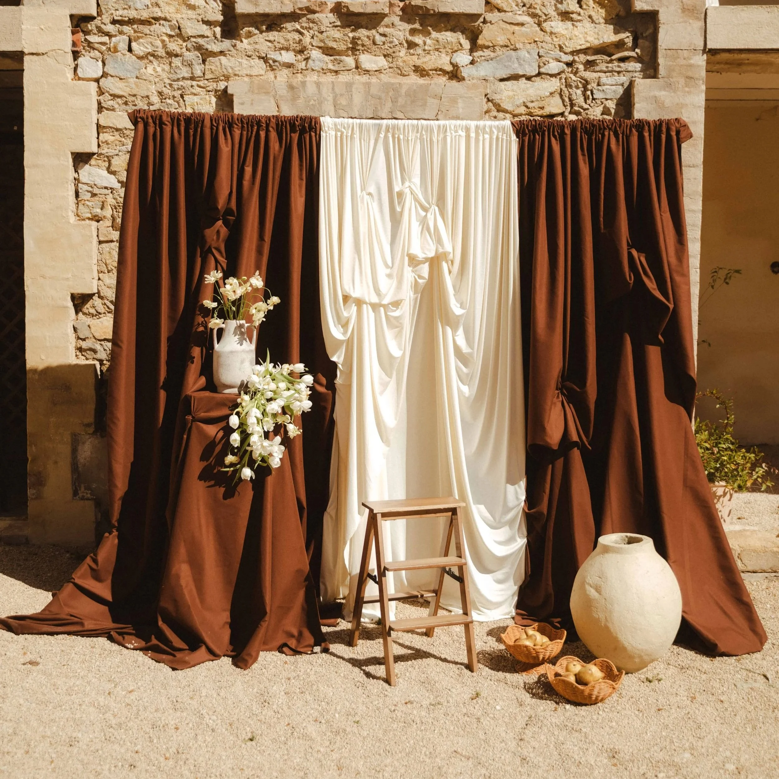Décor de scène antique avec des rideaux bruns et blancs, une table en bois, un vase blanc avec des fleurs blanches, une panière avec des pommes, et un fond en pierre ancienne.