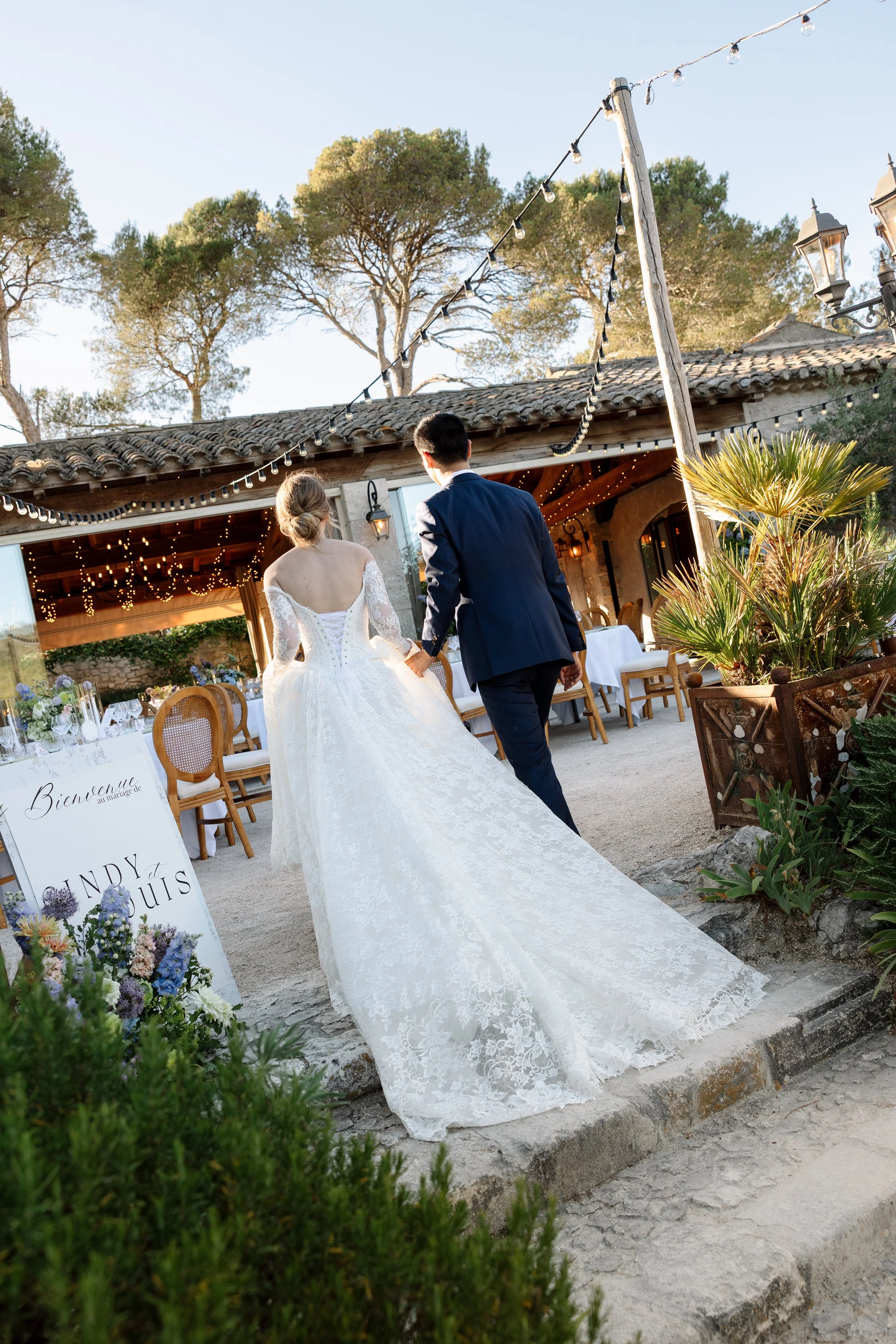 Un couple se tenant par la main lors d'un mariage en plein air devant une maison avec des lumières suspendues, entourée de plantes et d'arbres.