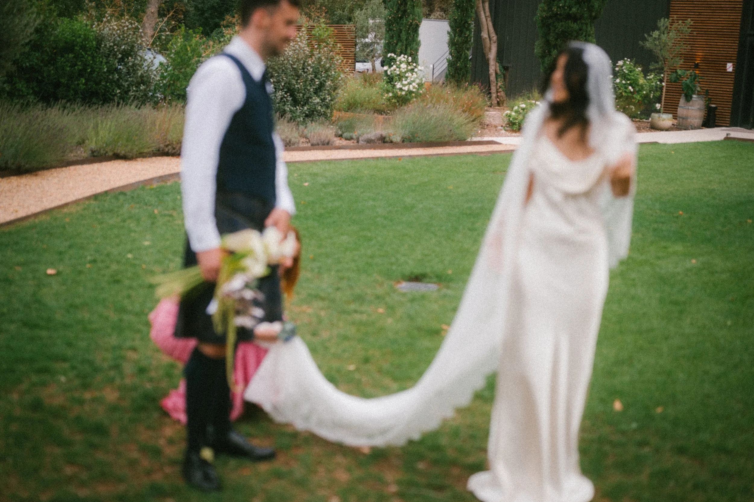 Un homme et une femme lors d'un mariage, l'homme tient un bouquet de fleurs, la femme porte une robe de mariée, ils se tiennent sur une pelouse verte avec des arbustes et des fleurs en arrière-plan