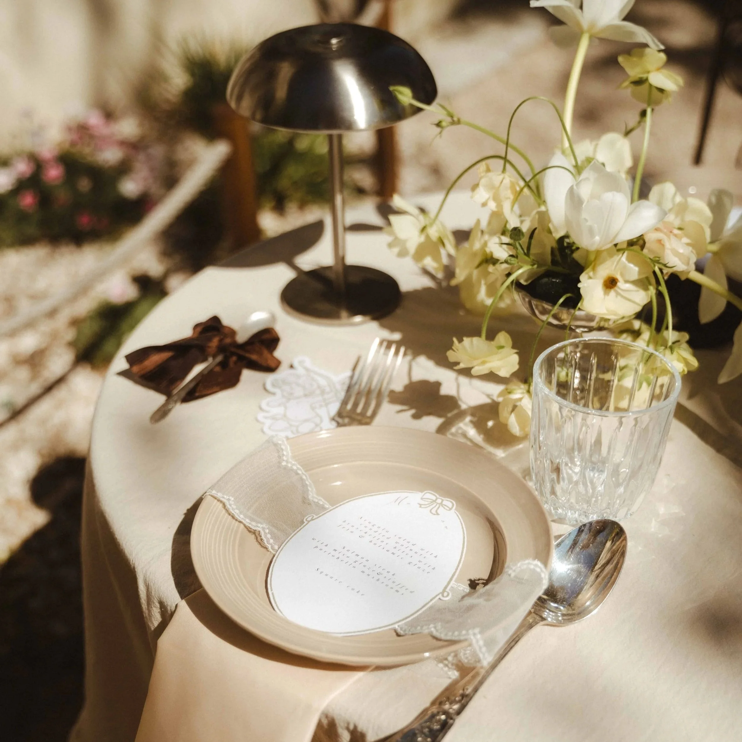 Une table avec une assiette blanche, un verre à pied en cristal, un bouquet de fleurs blanches, une petite boîte ronde avec un motif doré et une fourchette en vermeil.