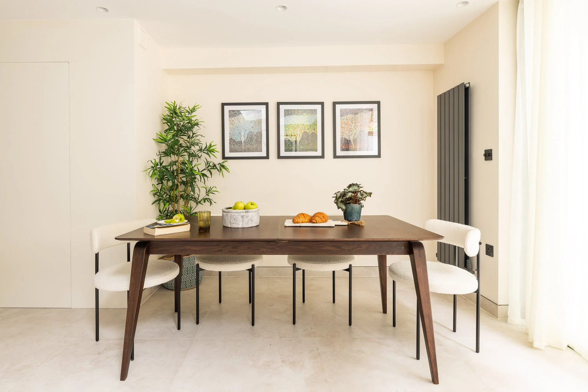 Dining room with a wooden table, four white upholstered chairs, two on each side, a large potted plant, three framed abstract art pieces on the wall, and natural light coming through a window with curtains.