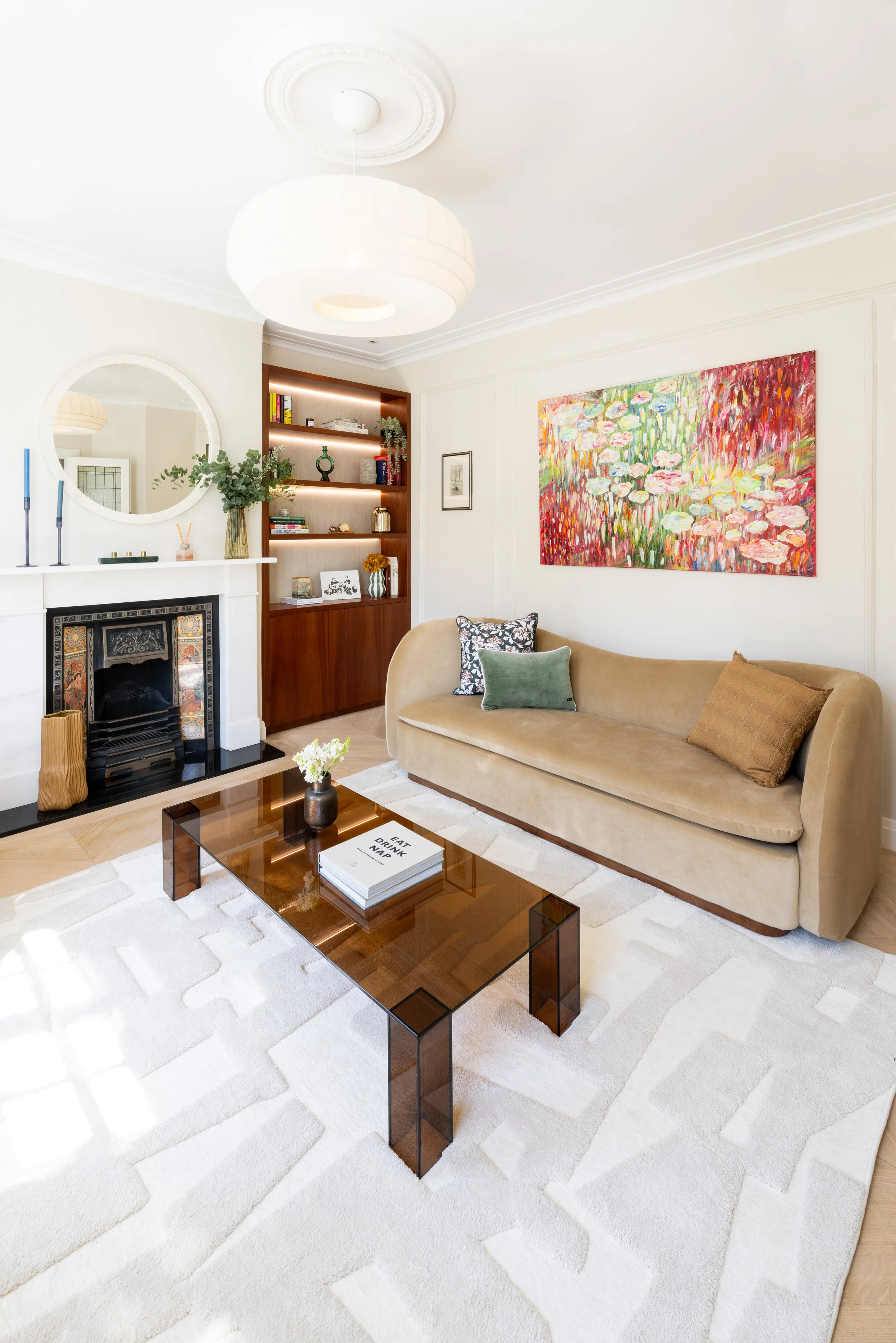 Living room with beige sofa, abstract painting, white rug, glass coffee table, fireplace, mirror, built-in wooden shelves with books and decorations, ceiling light fixture.