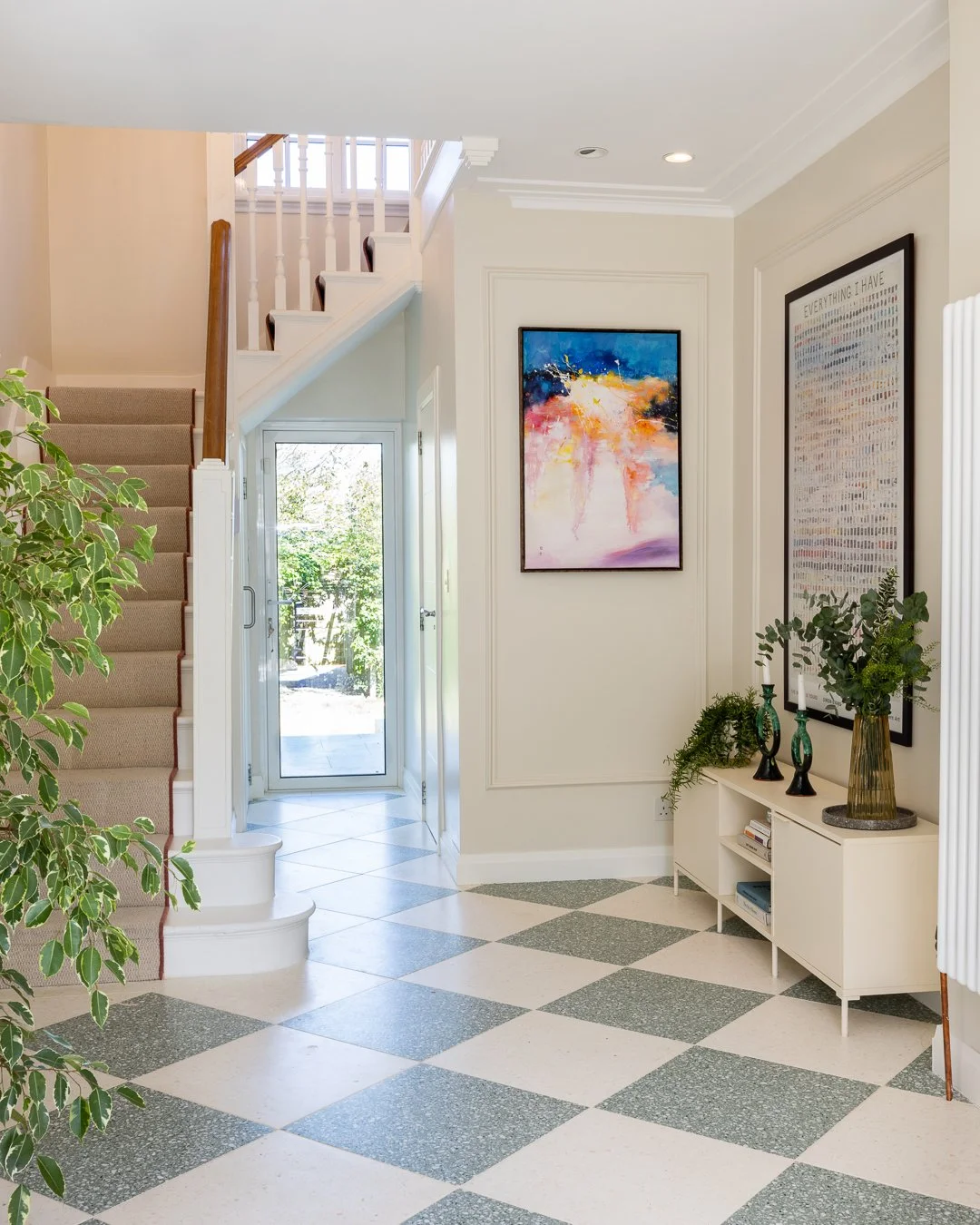Entryway of a house featuring a staircase with beige carpet, black and white checkered floor, a white console table with vases and plants, colorful abstract paintings on the walls, a glass door leading outside, and greenery visible through the door.