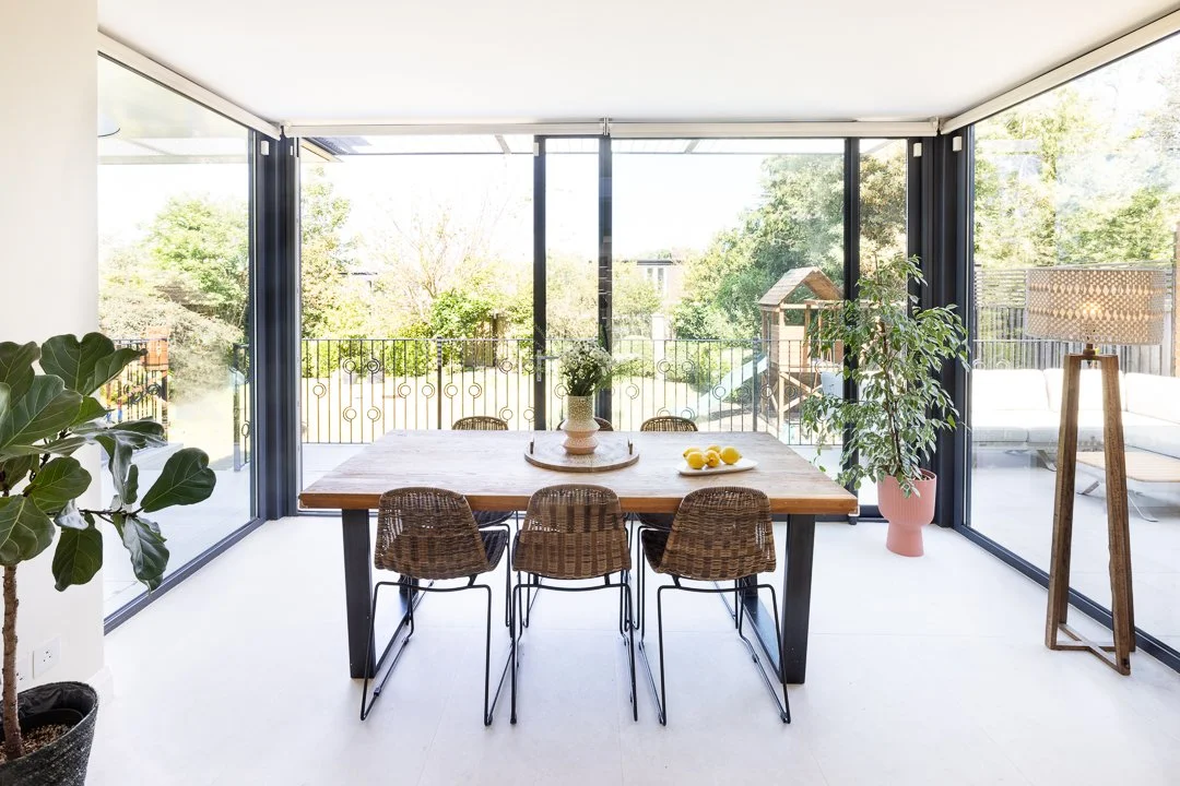 Modern dining room with large glass windows, wooden table, six wicker chairs, potted plants, and a tray of yellow lemons.