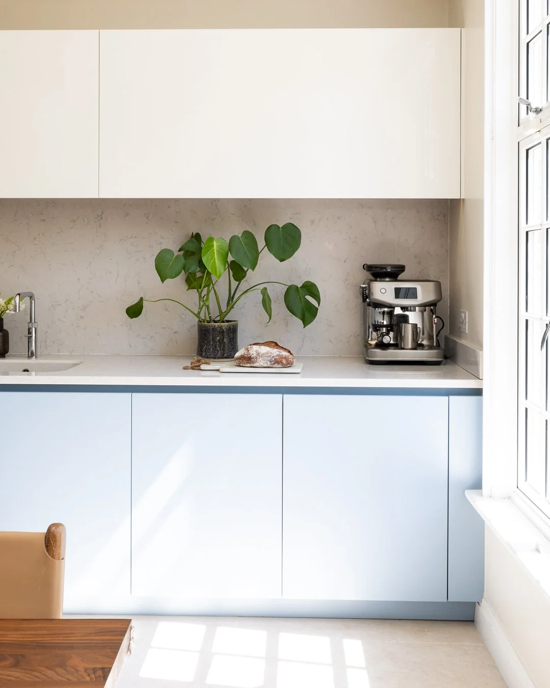 Minimalist kitchen with white cabinets, a potted plant, a loaf of bread, a coffee machine, and a window letting in natural light.