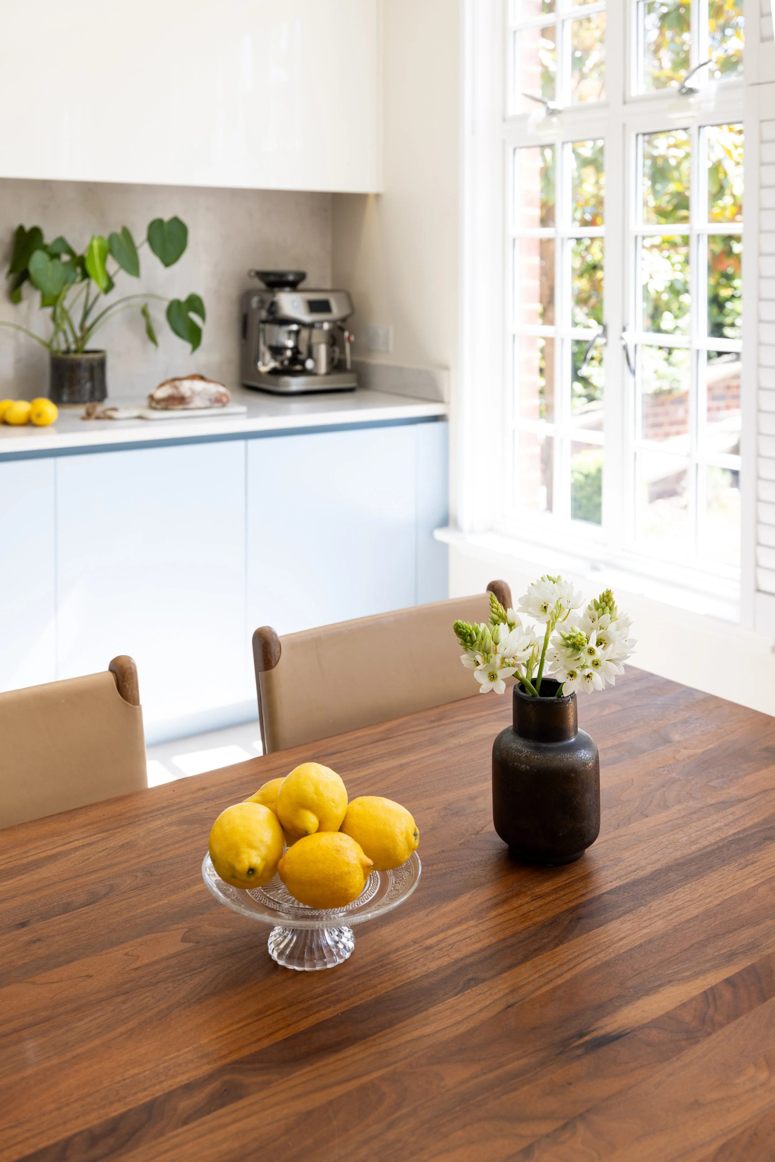 A wooden dining table with a black vase of white flowers and a glass plate of lemons.