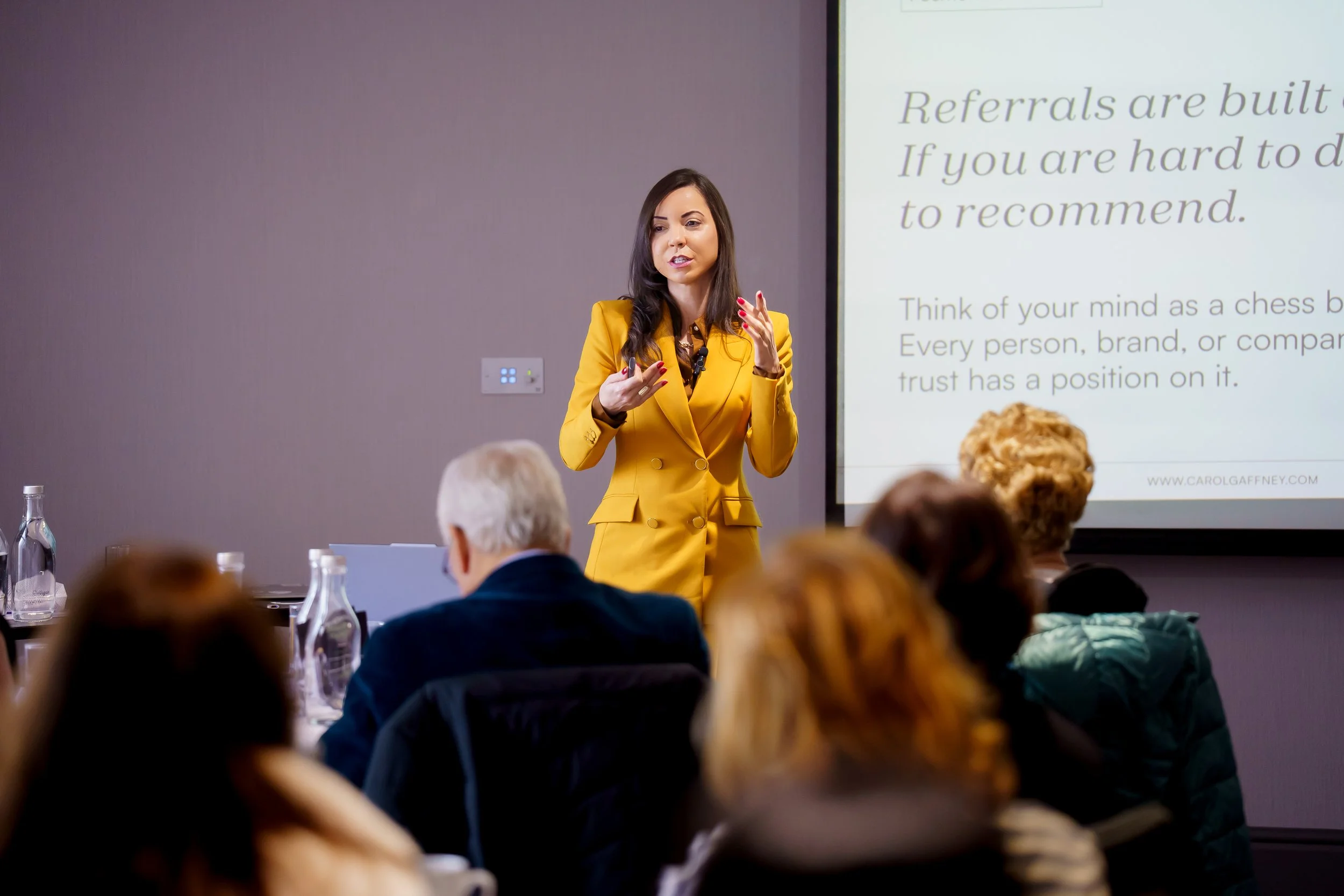 A woman in a yellow blazer giving a presentation in a conference room with a large screen displaying text. The audience is seated, listening attentively.