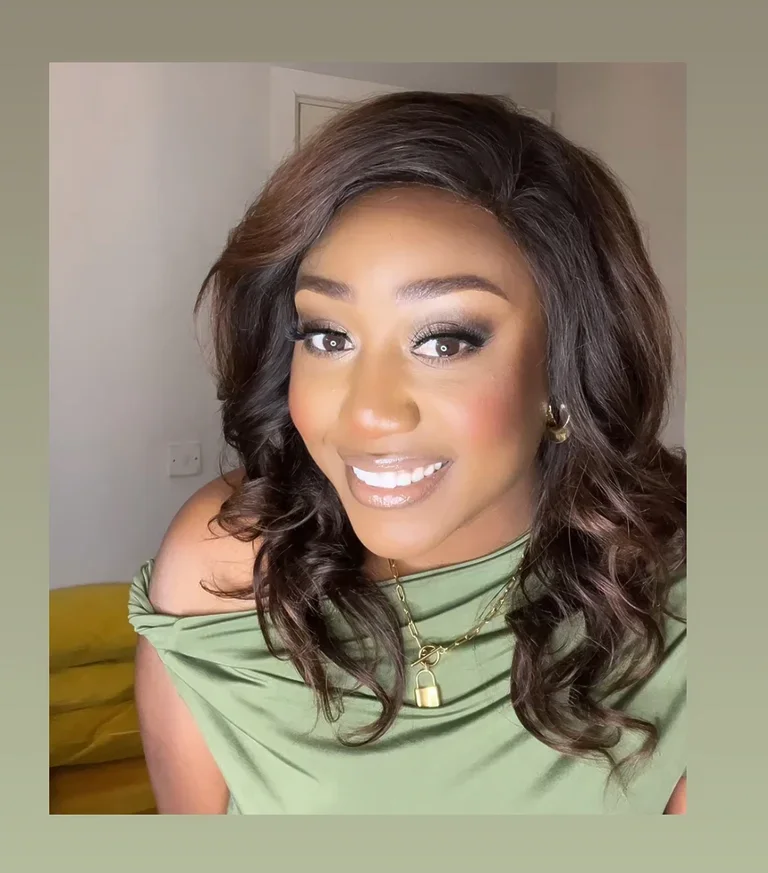 Close-up of a smiling woman with wavy dark hair, wearing makeup with shiny lips, gold jewelry, and an off-the-shoulder green top, indoors.