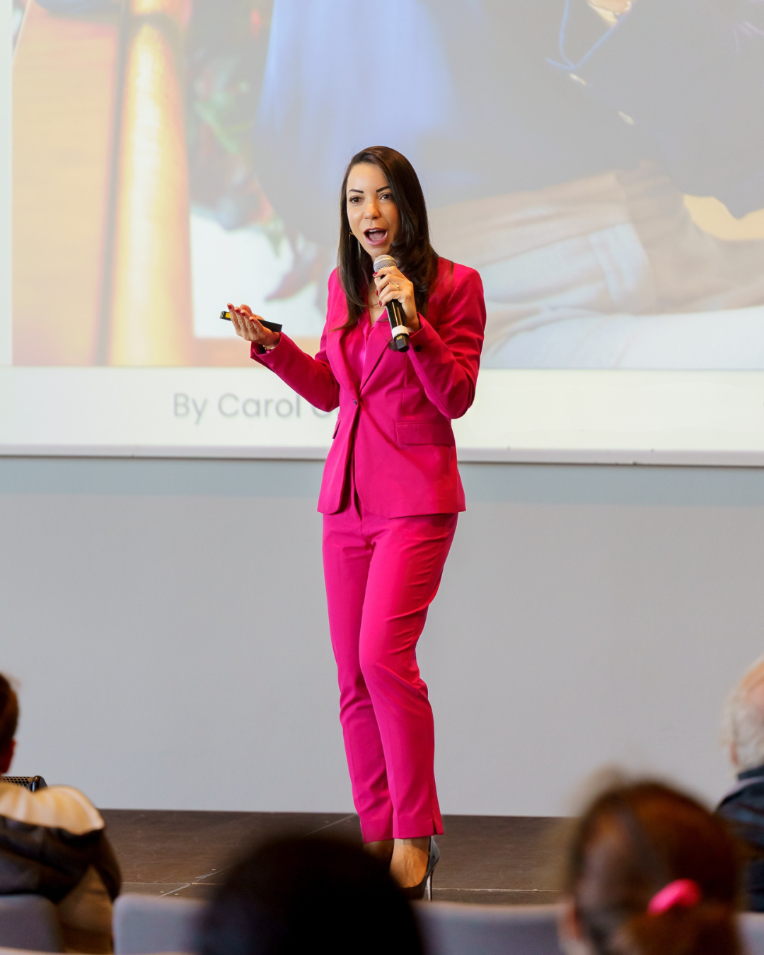 A woman in a bright pink suit giving a presentation on stage, holding a microphone in her right hand and a remote in her left, with a large screen behind her showing a slide.
