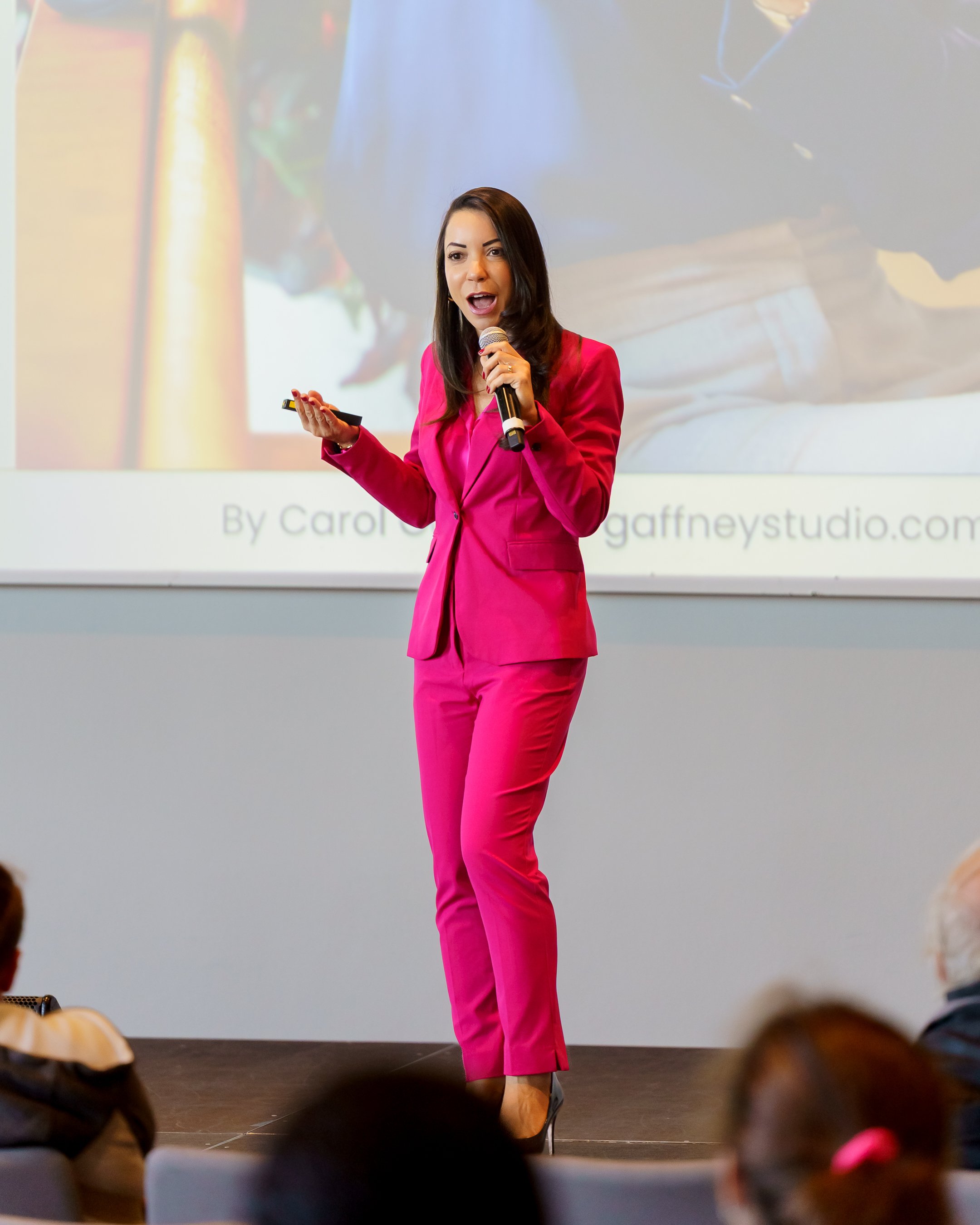 Woman in a bright pink suit giving a presentation, holding a microphone and a remote control in front of an audience.