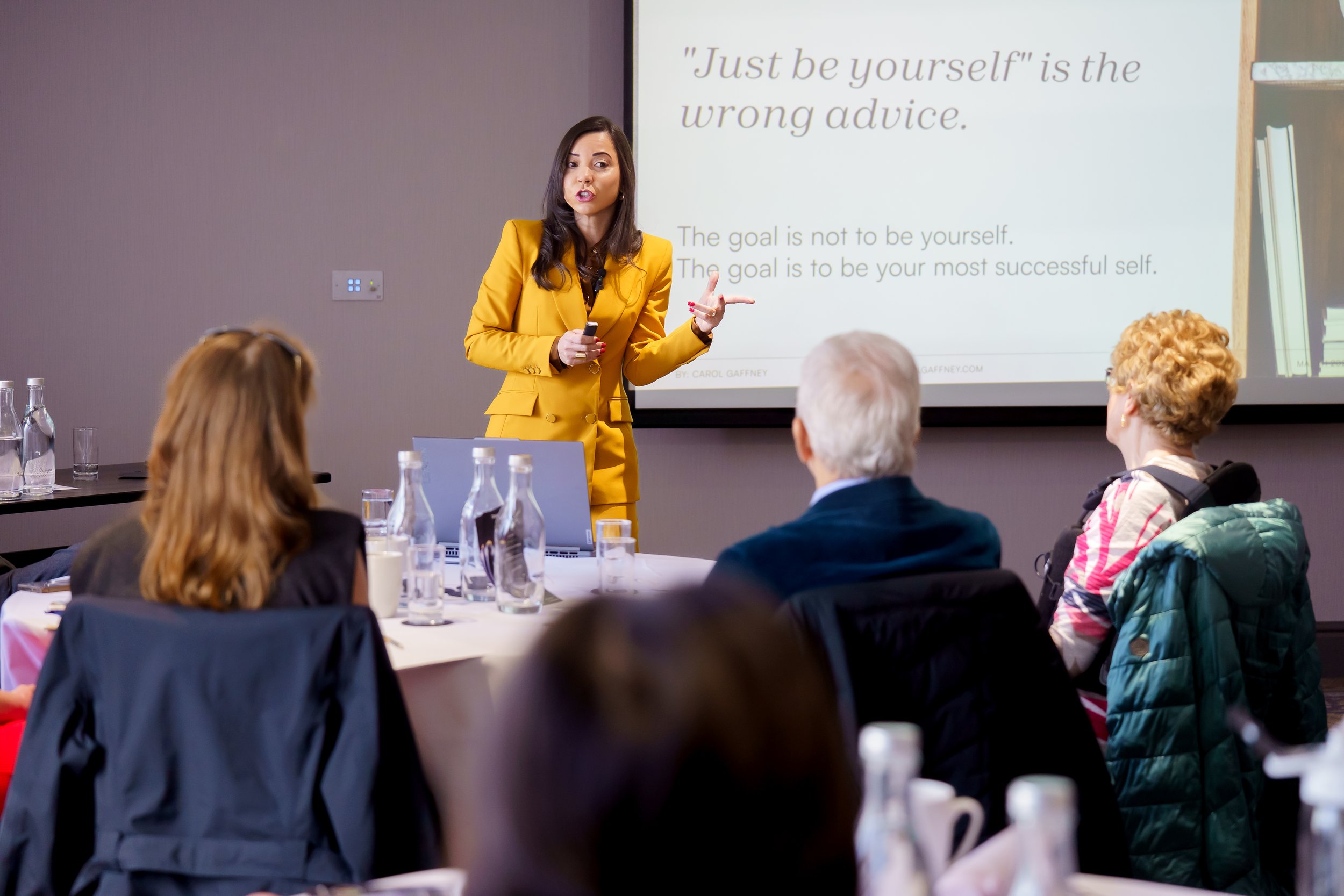 A woman in a yellow blazer giving a presentation to an audience in a conference room. She is standing in front of a screen with a quote that reads "Just be yourself" is the wrong advice. The audience is seated at a round table with bottles of water and glasses.