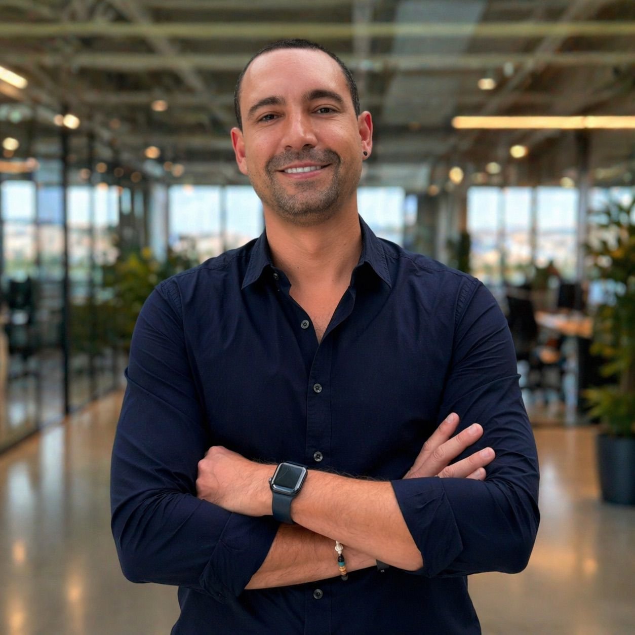 A man with short dark hair and a small beard, smiling with arms crossed, wearing a dark blue button-up shirt and a smartwatch, standing inside a modern office with large windows, plants, and blurred background.