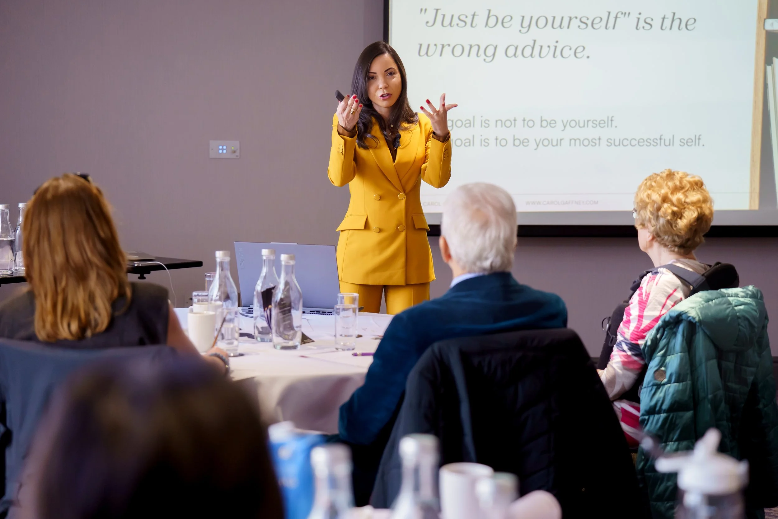 A woman in a yellow suit is giving a presentation to a group of people seated at a conference table. The presentation slide behind her reads, 'Just be yourself' is the wrong advice.