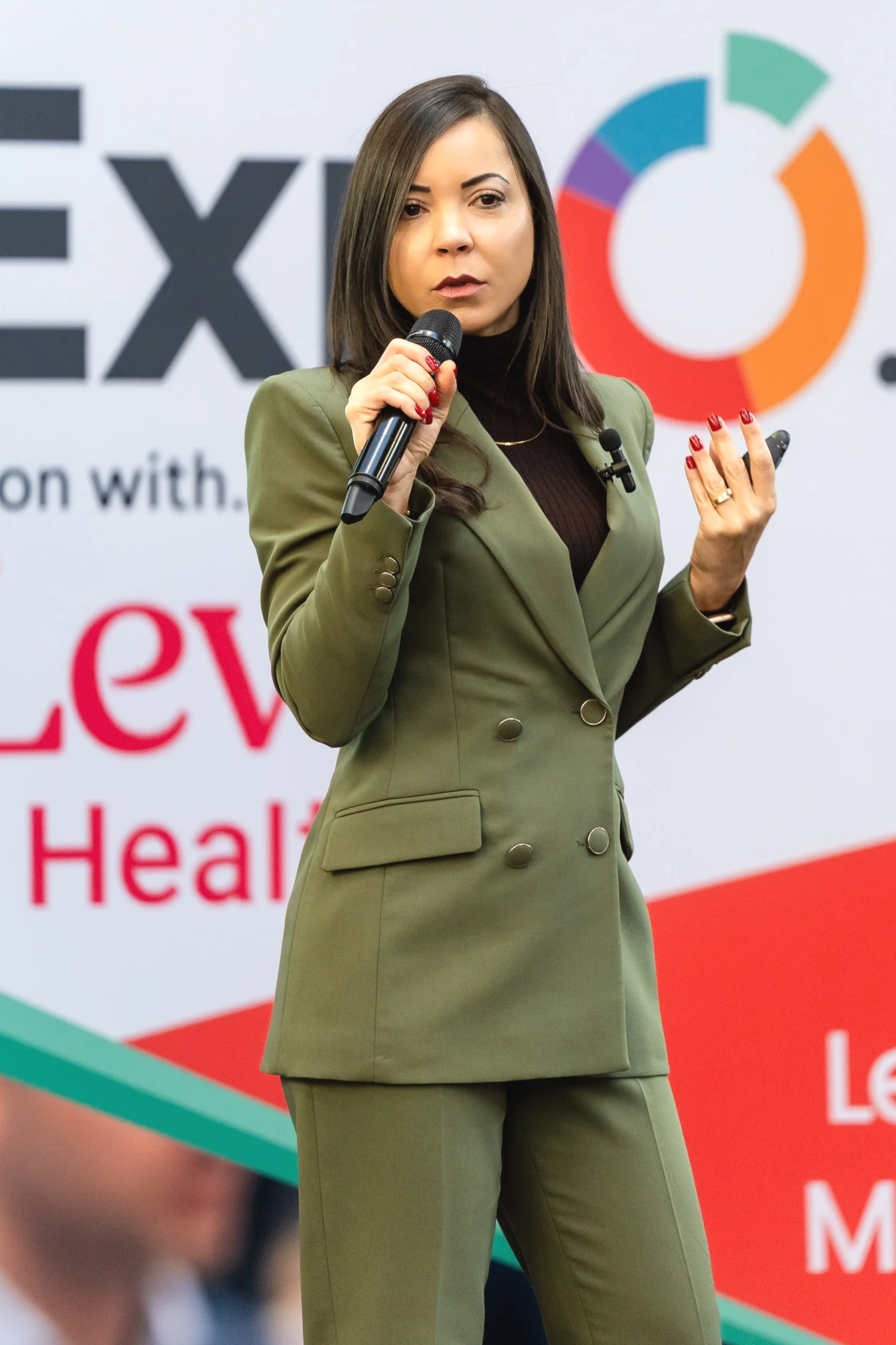 A woman in a green suit holding a microphone and giving a presentation in front of a colorful circular chart and a large sign with partially visible red and black text.