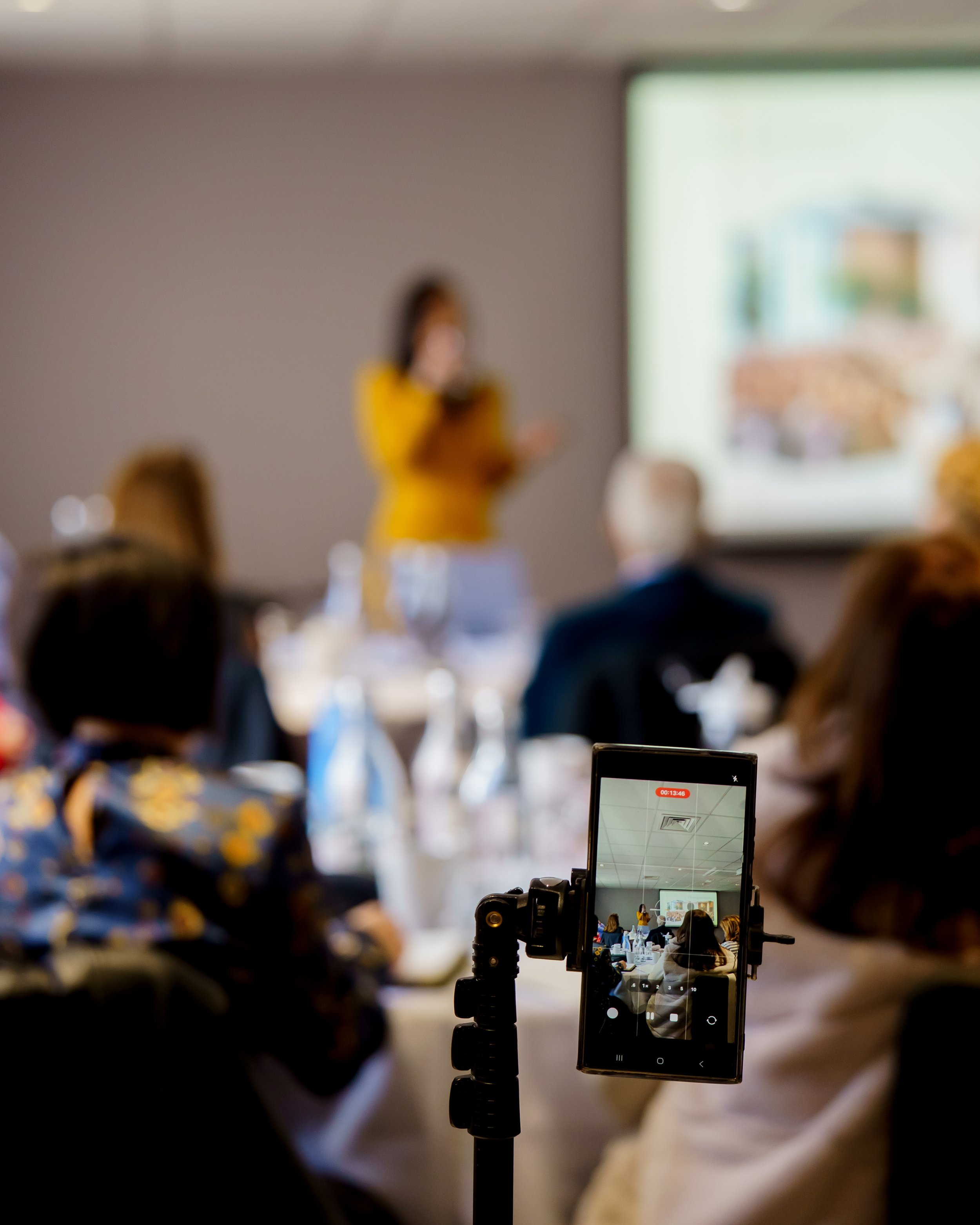 A blurred image of a woman in a yellow top giving a presentation in a conference room, with an audience seated at tables. A smartphone on a tripod is capturing the event, showing the woman, the presentation, and the room on its screen.