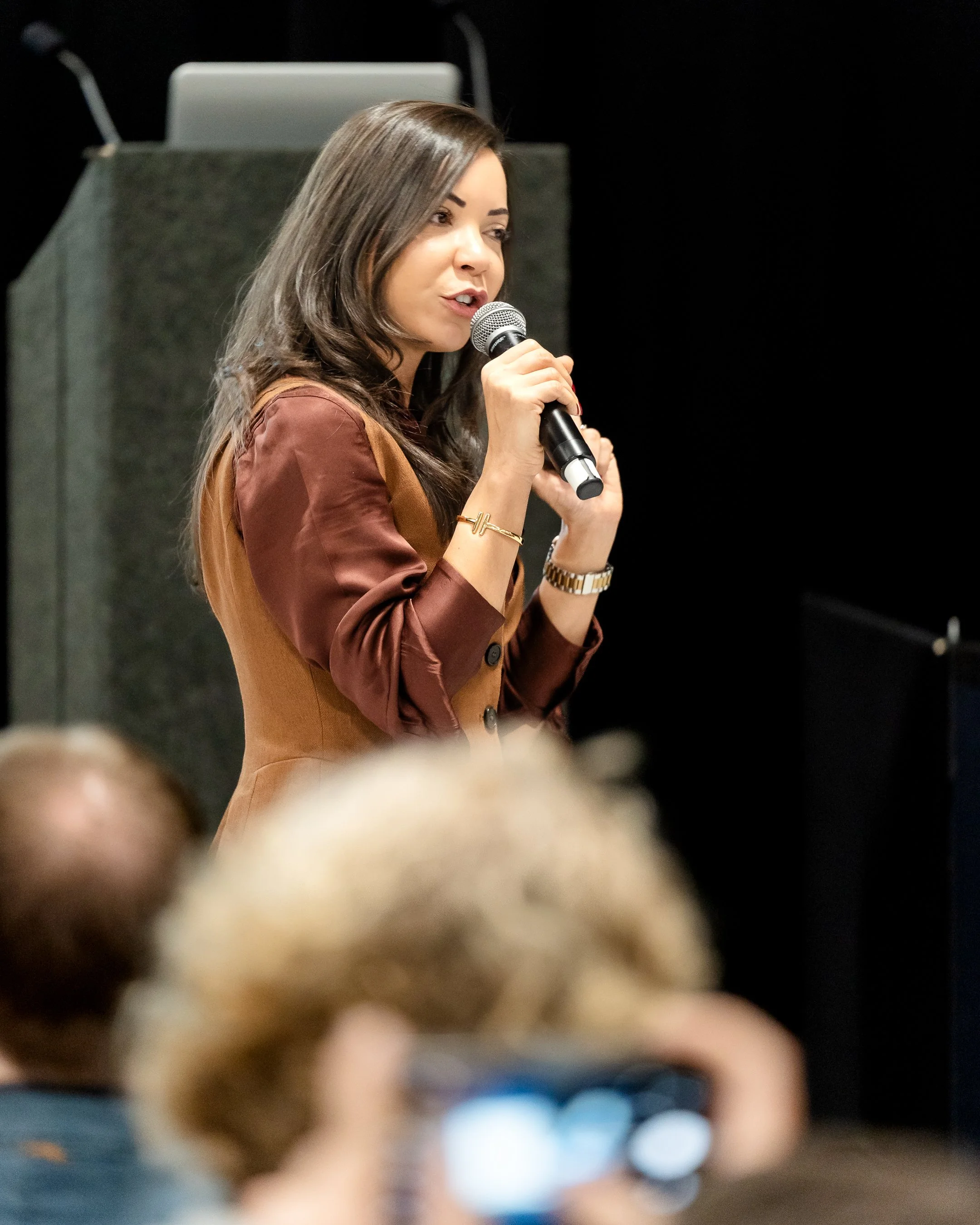A woman with long brown hair, wearing a brown blazer, speaking into a microphone during a presentation or speech in front of an audience.