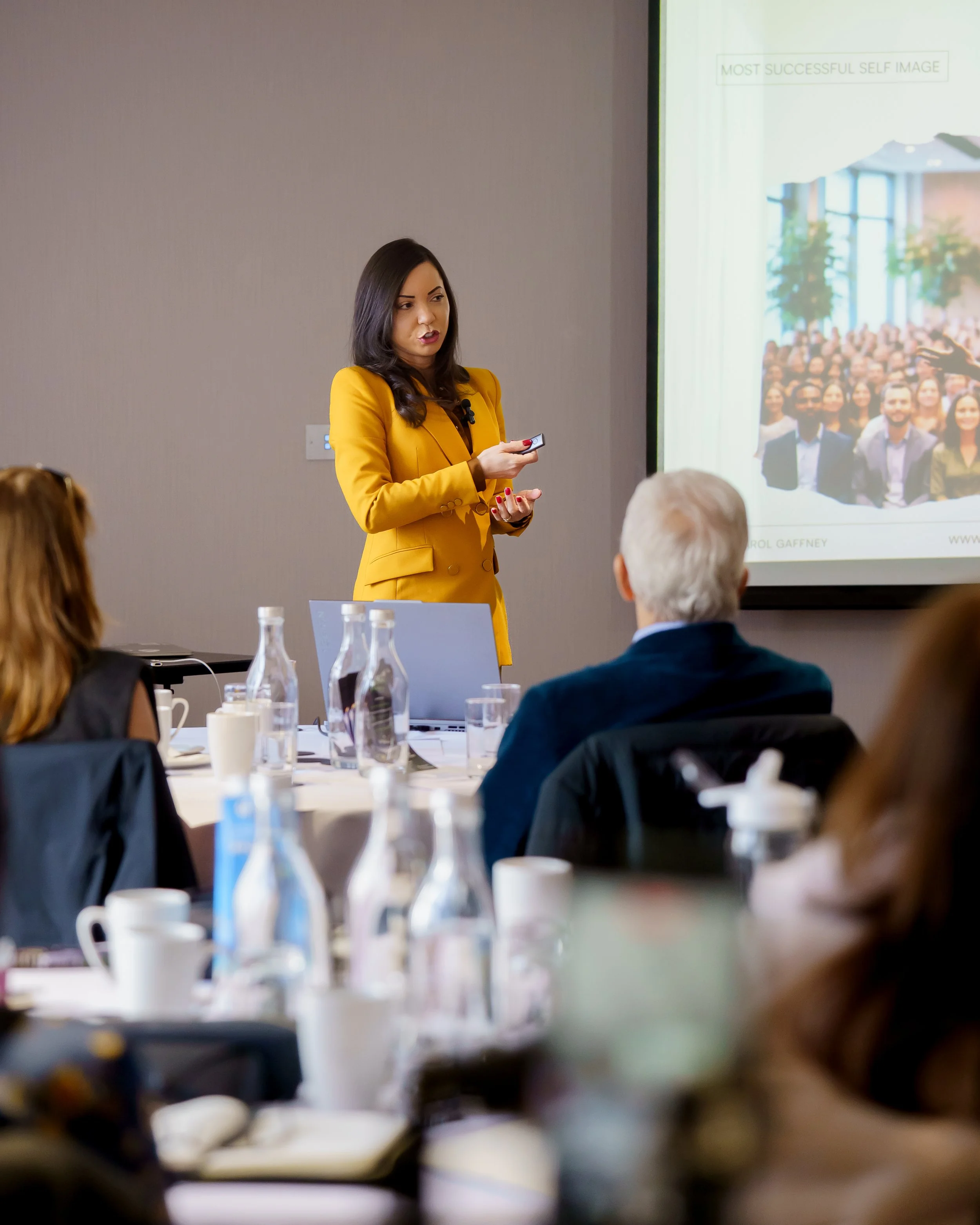 A woman in a yellow blazer is giving a presentation in a conference room. She is holding a remote control and standing near a large screen displaying a slide titled 'Most Successful Self Image'. There are seated attendees, some with their backs to the camera, in front of her, and bottles and cups on the table.