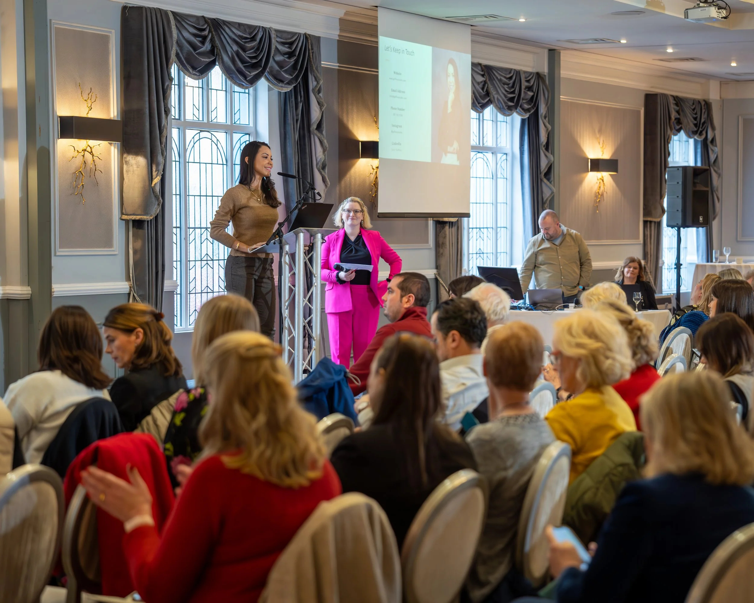 A woman giving a presentation at a conference with an audience seated in front of her, a speaker on stage with pink suit, and a man managing audio equipment in a spacious, elegant room.