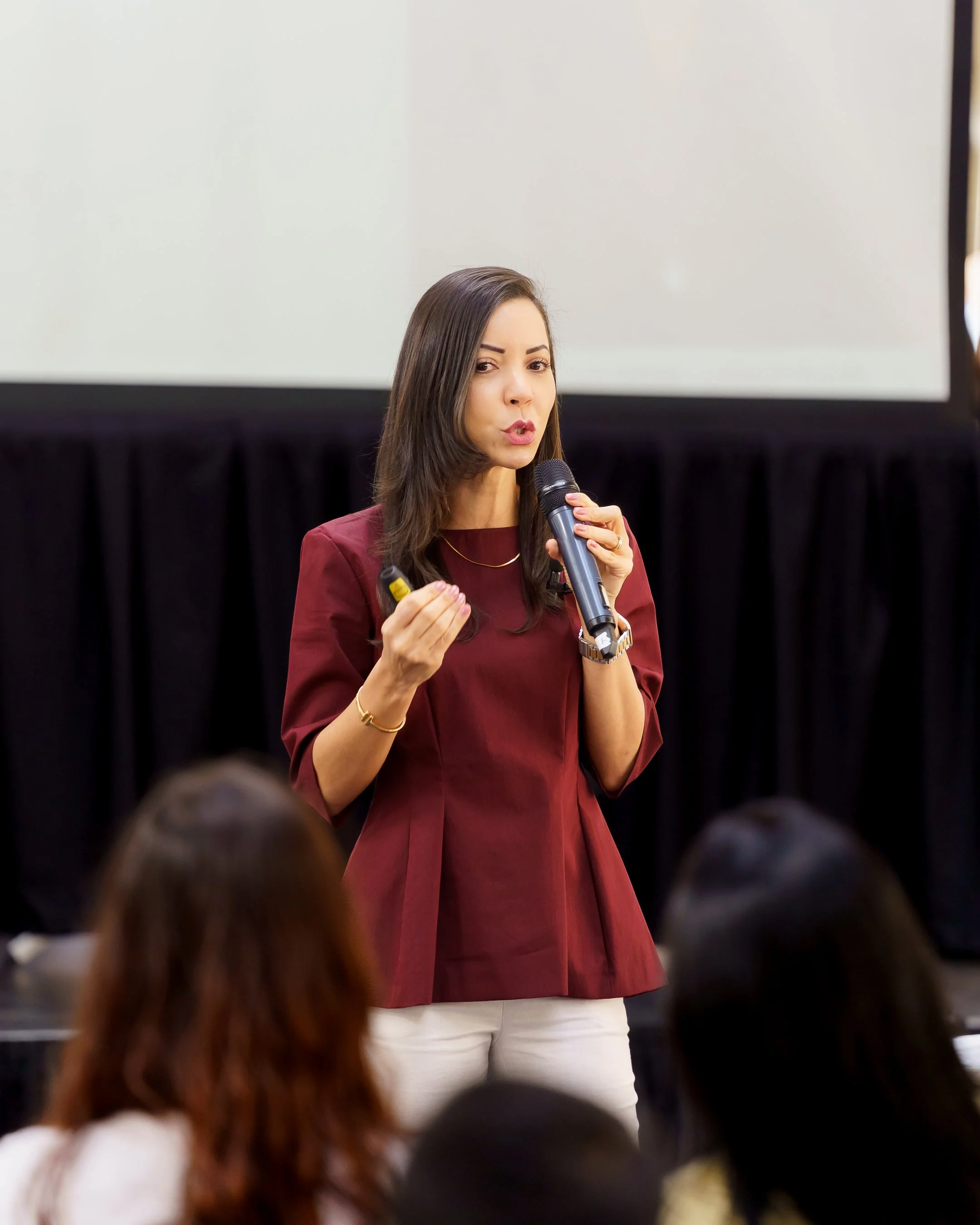 Woman giving a presentation, holding a microphone and a remote, in front of an audience.