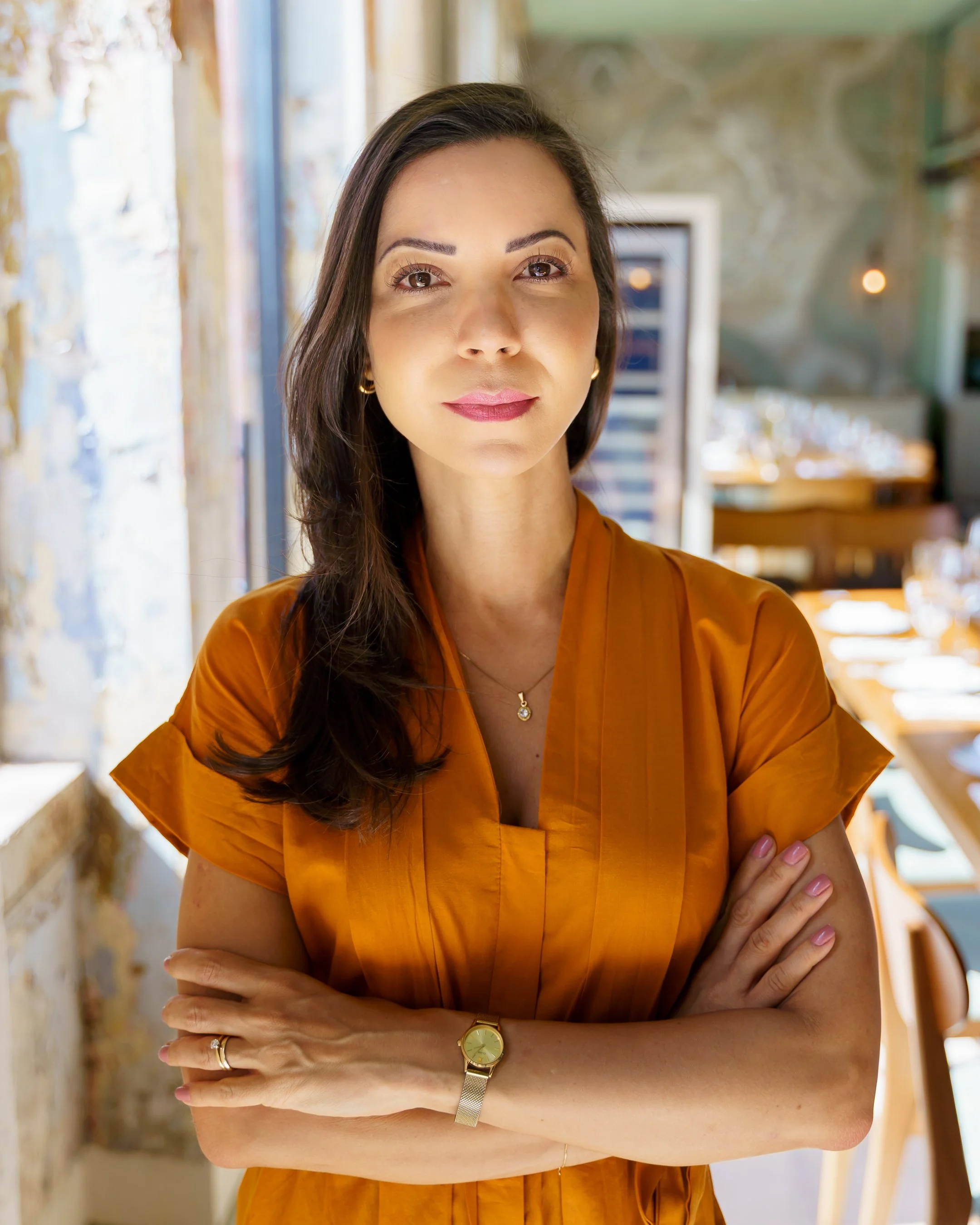 A woman with medium-length dark hair, wearing a mustard-colored top and gold jewelry, stands with her arms crossed in a sunlit interior space with textured walls and wooden furniture.
