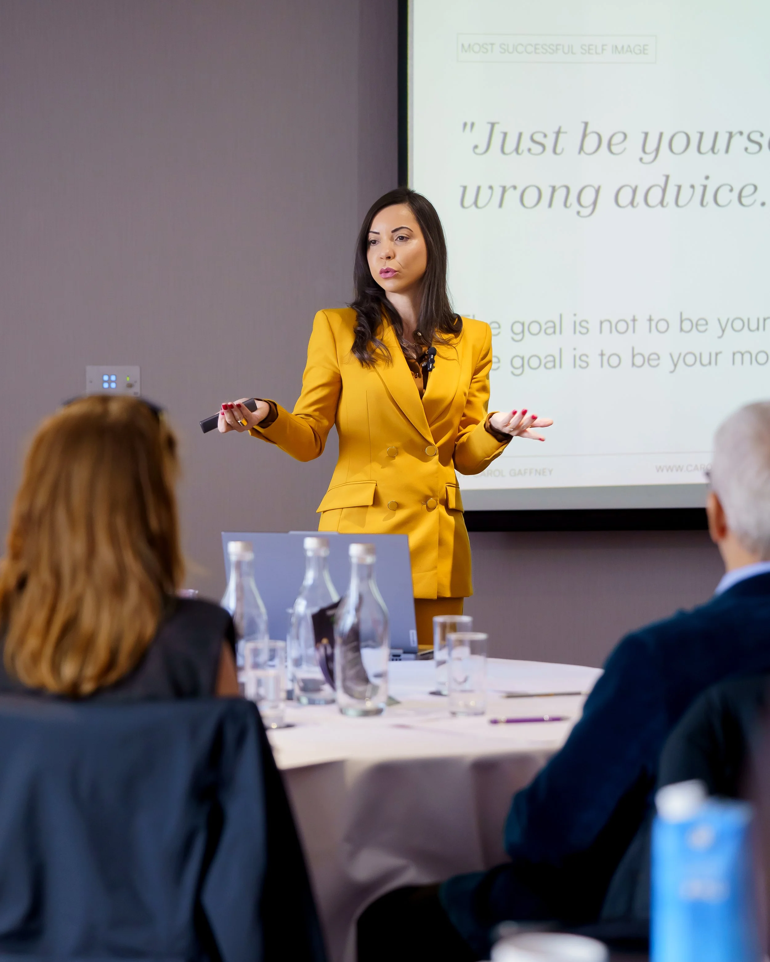 Woman in yellow suit giving a presentation in a conference room, with a slide in the background that says 'Just be yourself wrong advice'.