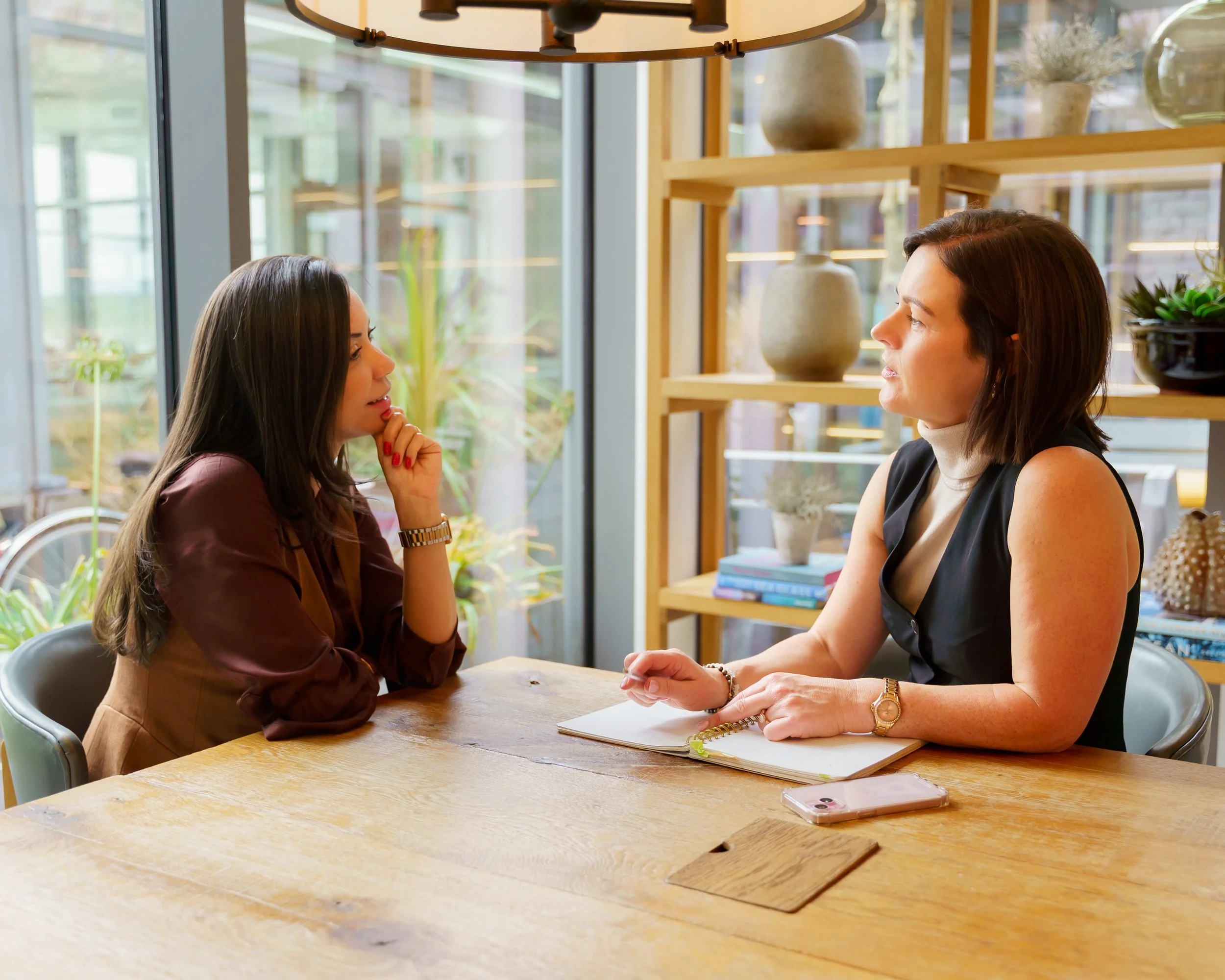 Two women sit at a wooden table in a modern, well-lit cafe, engaged in a serious conversation. The woman on the left has long dark hair and is wearing a brown blouse, resting her chin on her hand. The woman on the right has shoulder-length dark hair, is wearing a black sleeveless top, and has a notebook and a smartphone in front of her. Behind them is a shelf with decorative vases and plants, and large windows let in natural light.