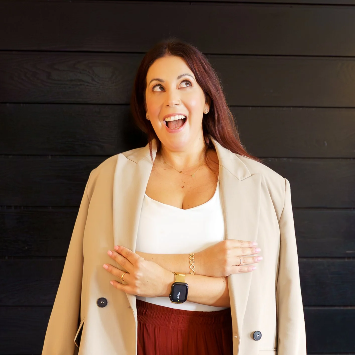 A woman with red hair smiling and looking up, wearing a beige blazer over a white top, a wristwatch, and a gold bracelet, standing against a dark wooden wall.