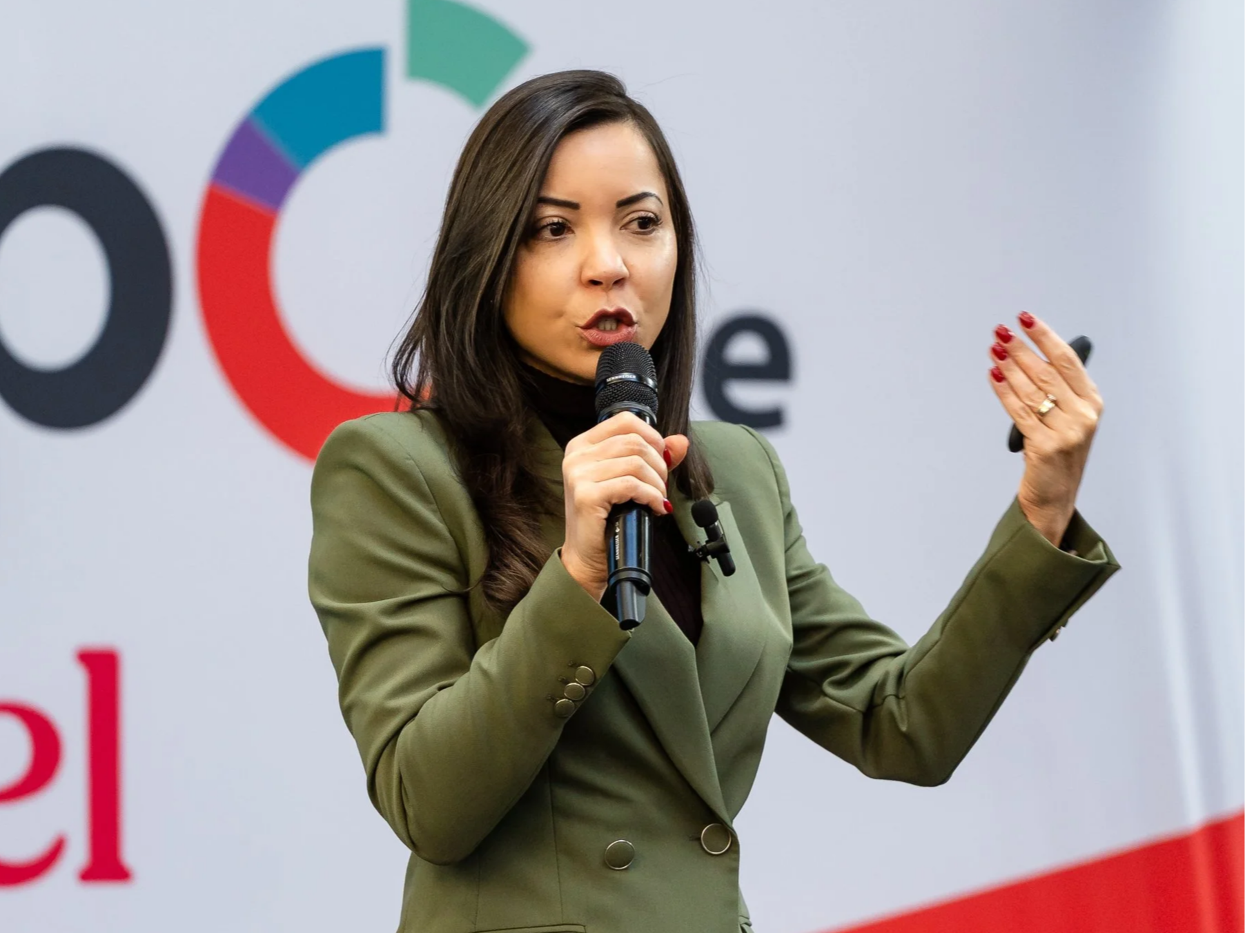 A woman with dark hair, wearing an olive green suit, speaking into a microphone at an event with a colorful logo in the background.