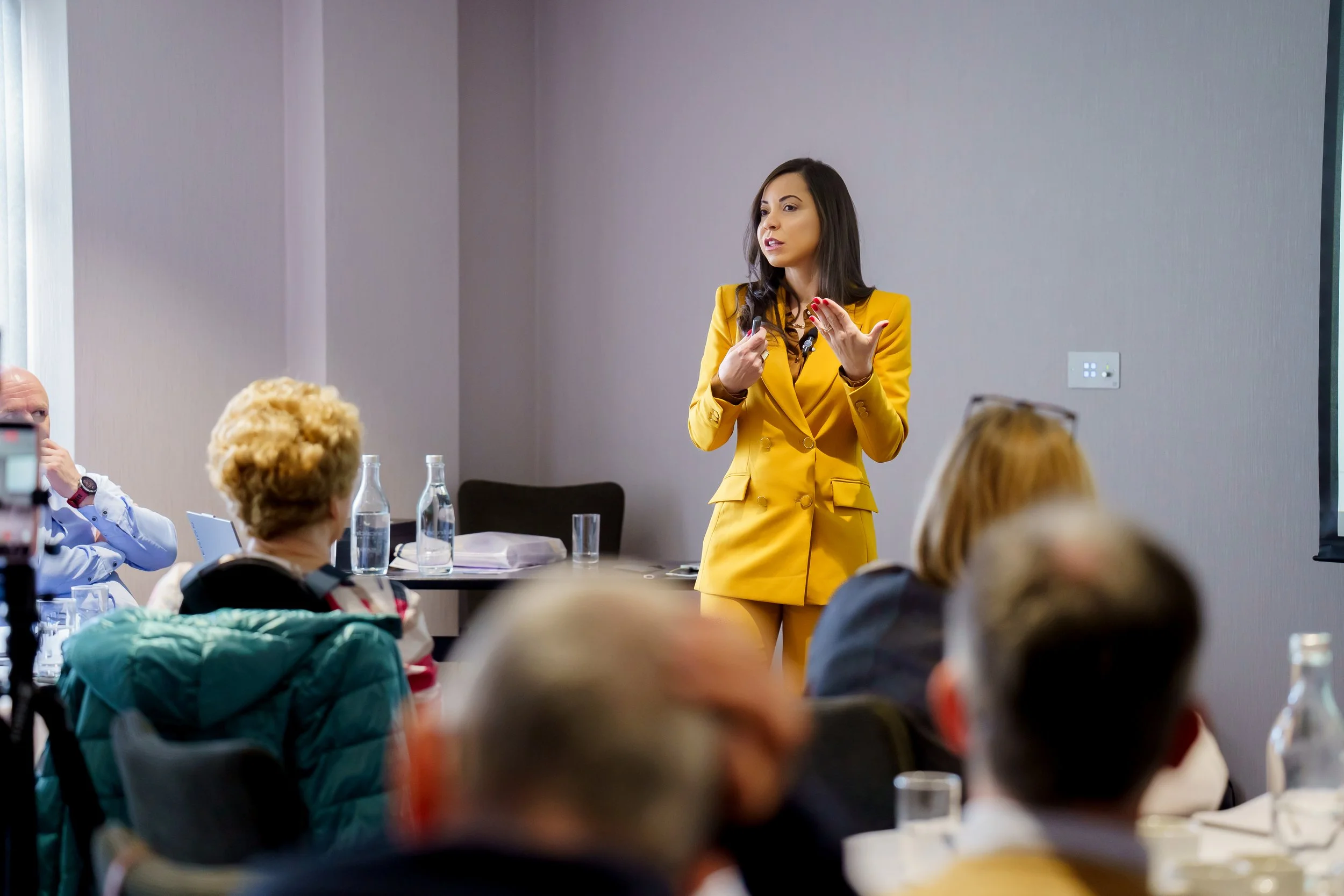 A woman in a yellow suit is giving a presentation to a group of people in a conference room.