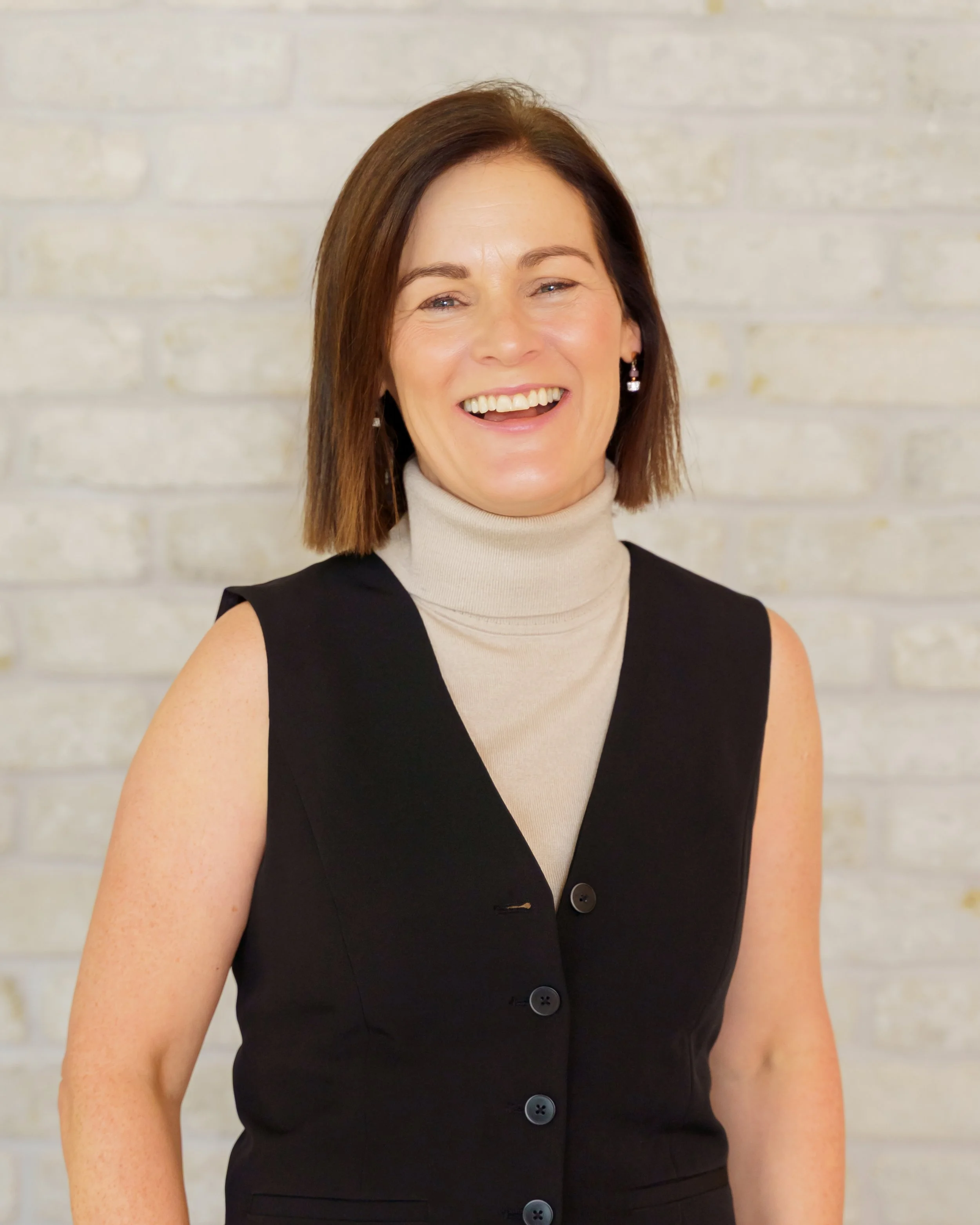 A woman with shoulder-length brown hair smiling, wearing a beige turtleneck and a sleeveless black vest, standing against a light-colored brick wall.
