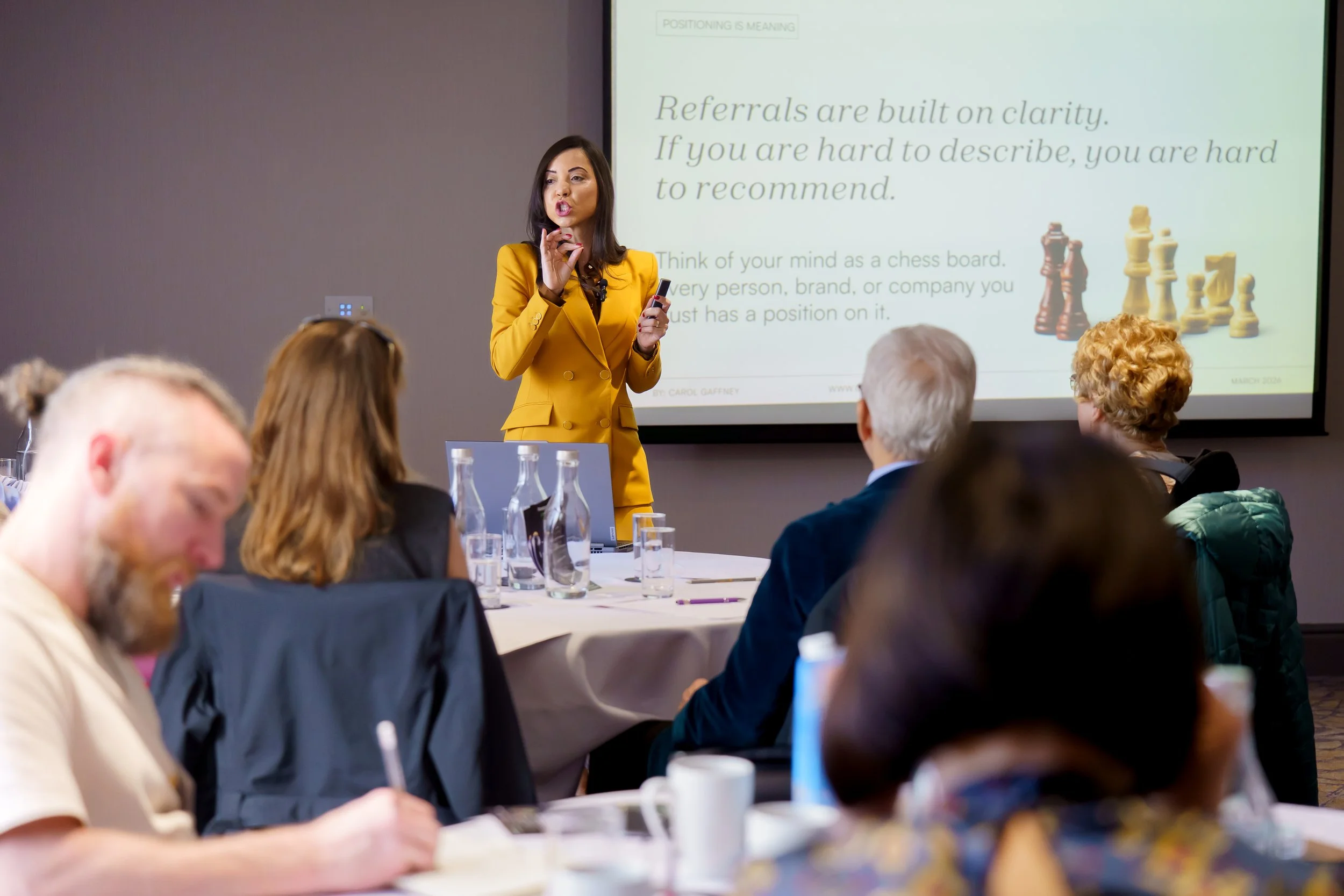 A woman in a yellow suit giving a presentation in a conference room with several attendees sitting at tables. A large screen behind her displays a slide about brand positioning and referrals.