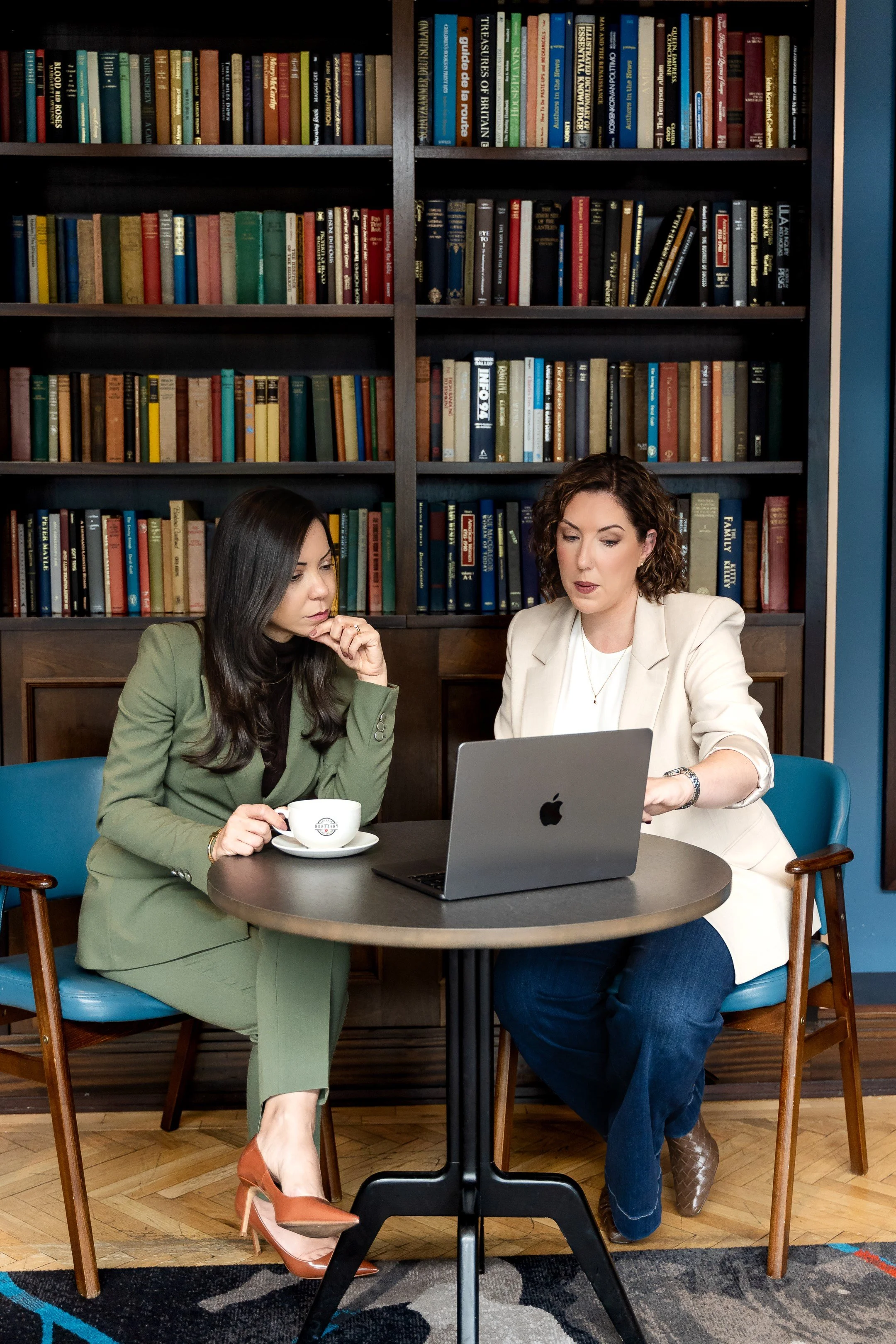 Two women sitting at a round table in front of a laptop and a bookshelf filled with books. One woman has long dark hair, is dressed in a green suit, and is holding a coffee cup. The other woman has curly brown hair, is dressed in a light blazer, and is gesturing towards the laptop.