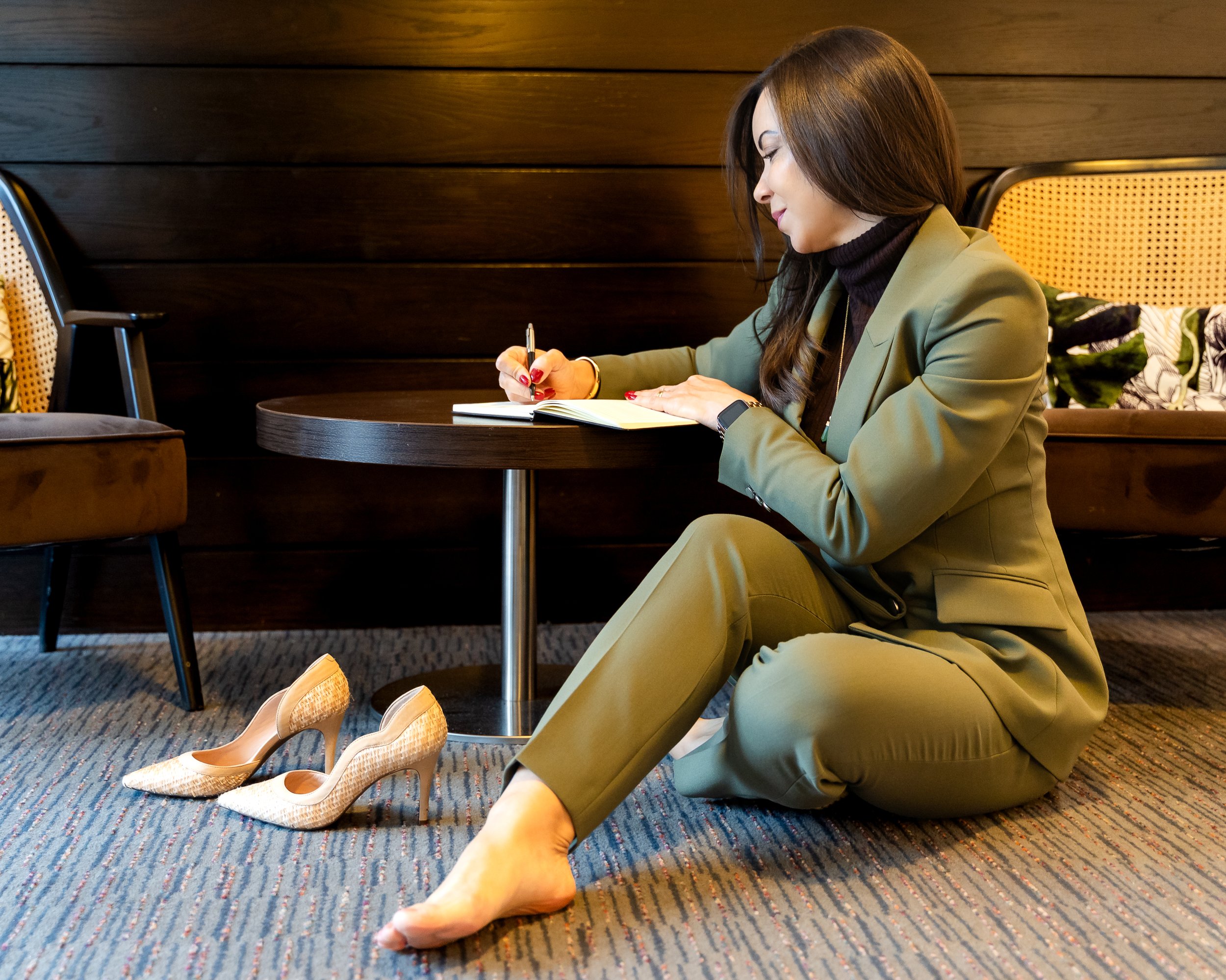Woman in a green suit sitting on the floor in a room, writing in a notebook on a small round table, with a pair of high heels on the carpet nearby.