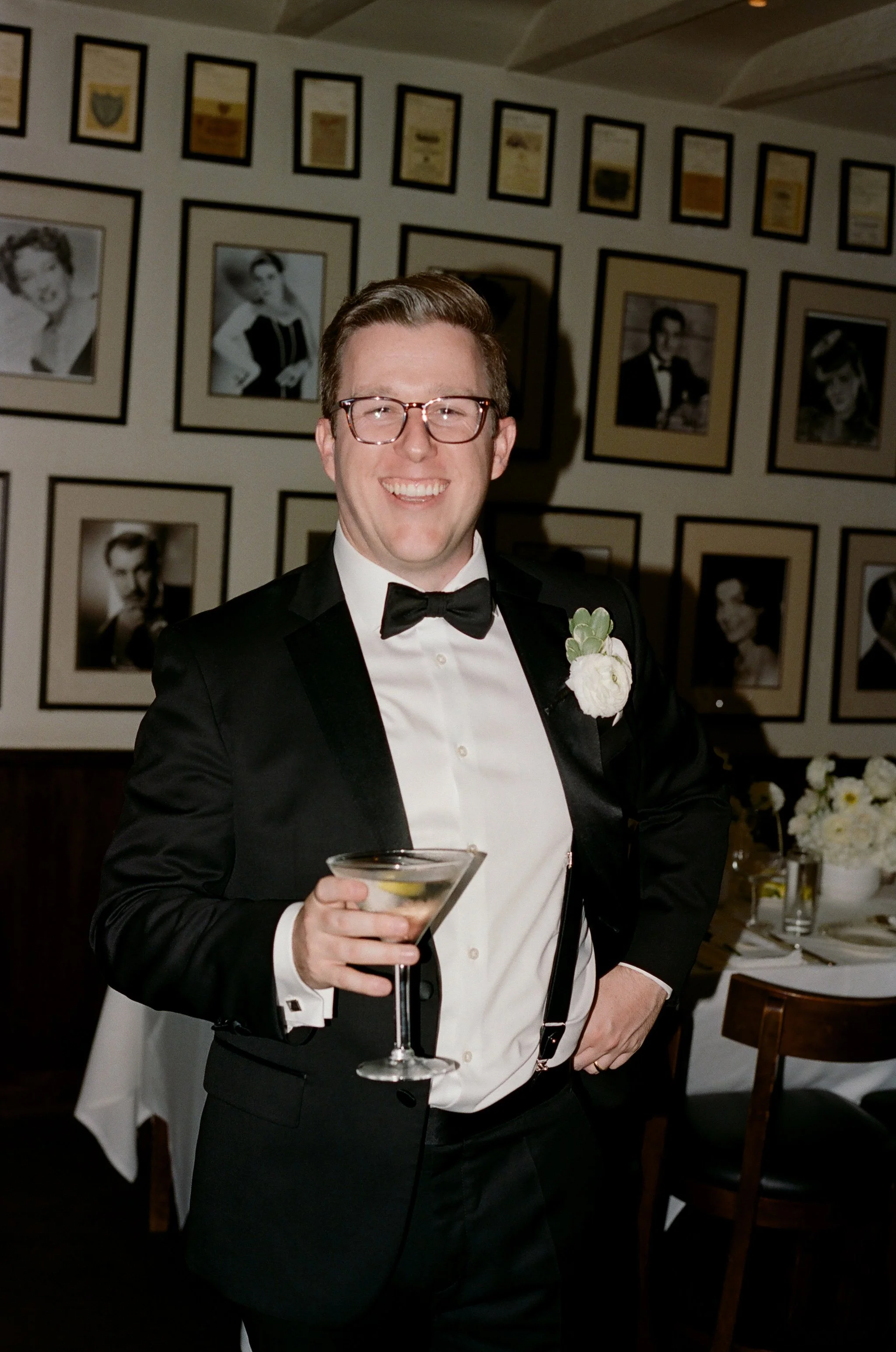 A man in a tuxedo with glasses, holding a martini glass, smiling at a formal event with framed black-and-white photographs on the wall behind him.