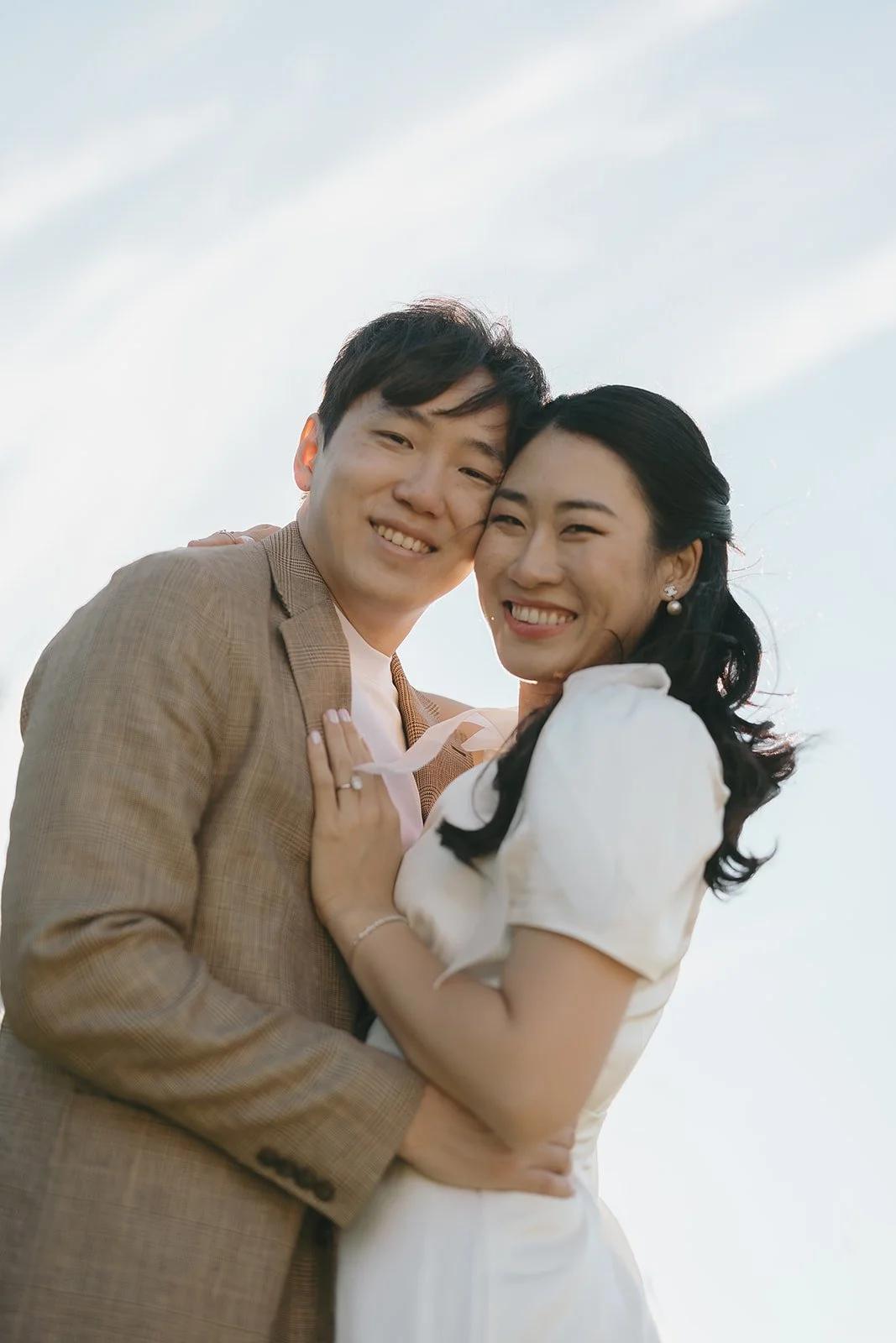 A smiling couple embracing outdoors against a light sky.