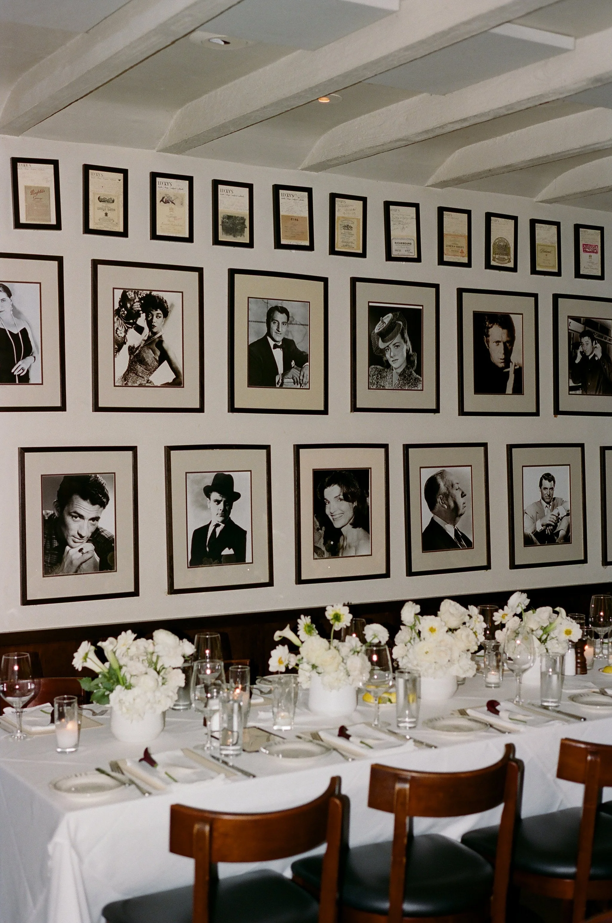 A dining table set for a formal meal with white tablecloth, glassware, plates, and floral centerpieces, against a wall decorated with framed black-and-white portraits of celebrities.
