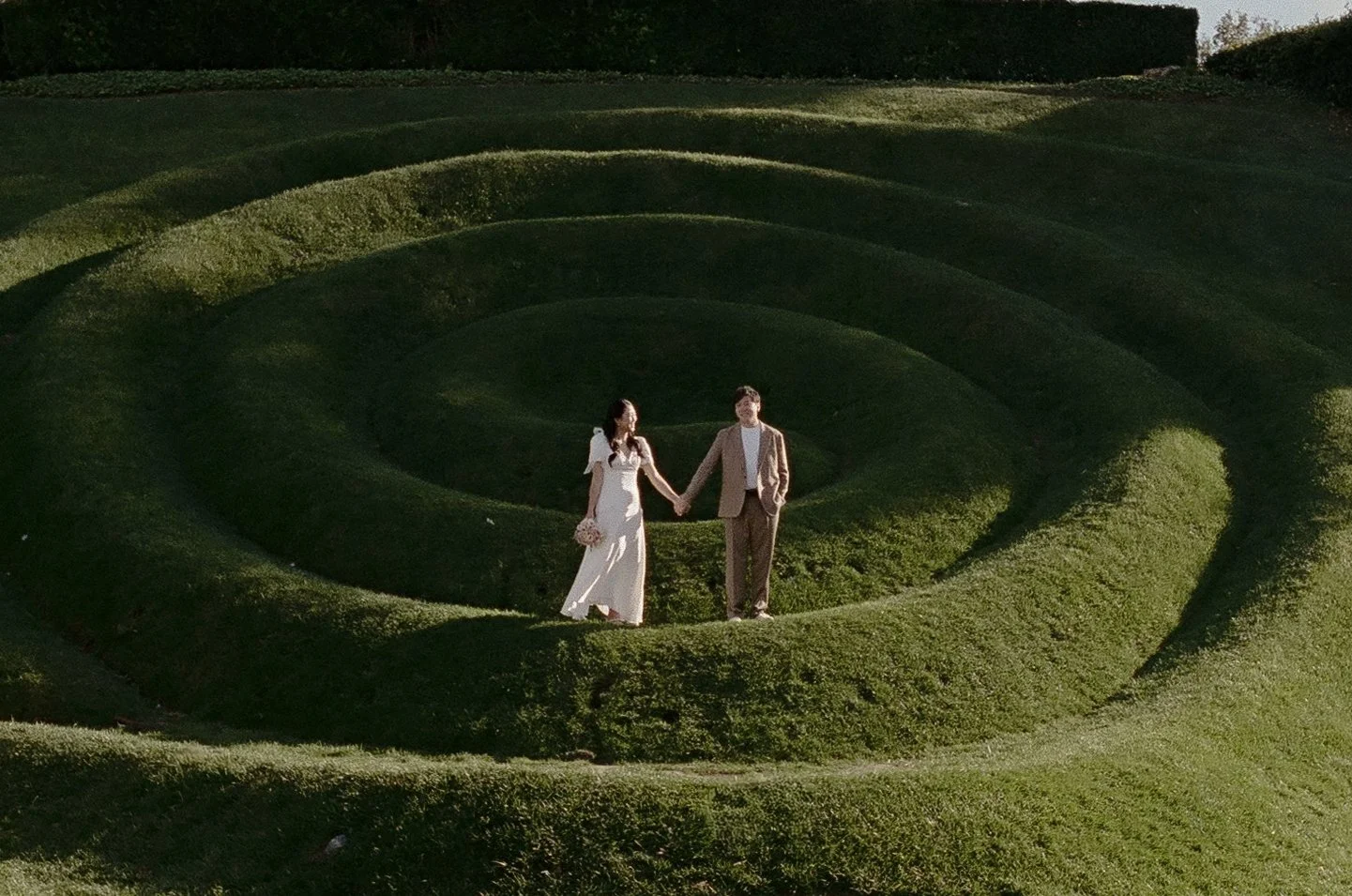 A couple walking hand in hand through a large, spiraling hedge maze in the daytime.