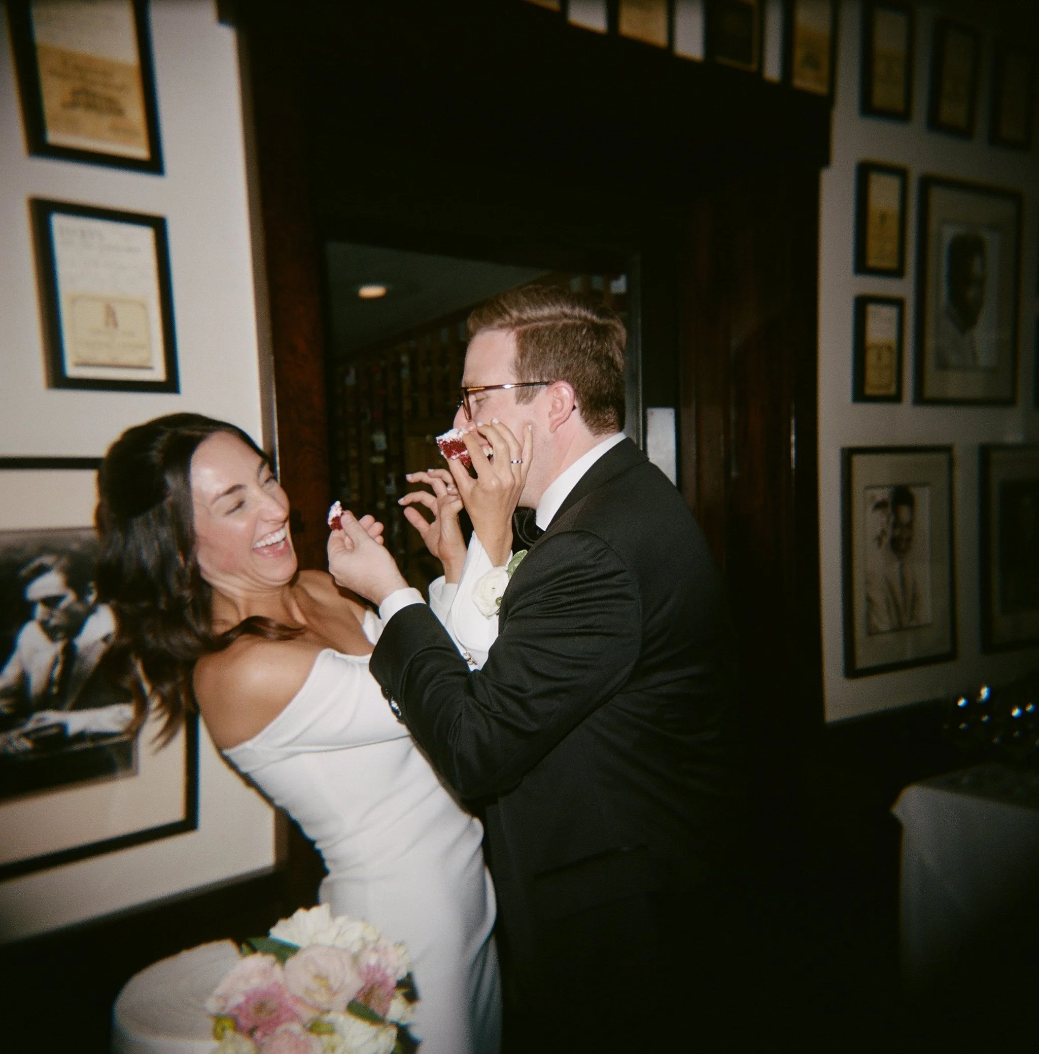 Bride and groom sharing a playful moment at their wedding reception, with the groom feedings cake to the bride in a room decorated with framed artwork on the walls.